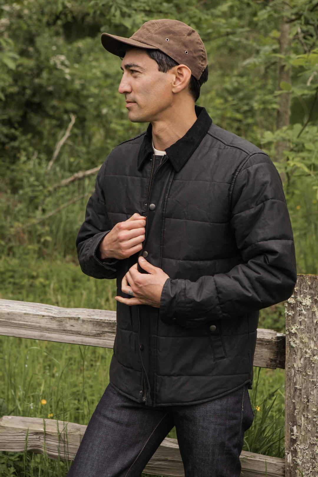 A man wearing a brown cap and Bridge & Burn's Holman Quilted Jacket in Black Waxed Canvas stands outdoors by a wooden fence, surrounded by green foliage, looking to the side with his hands on his jacket.