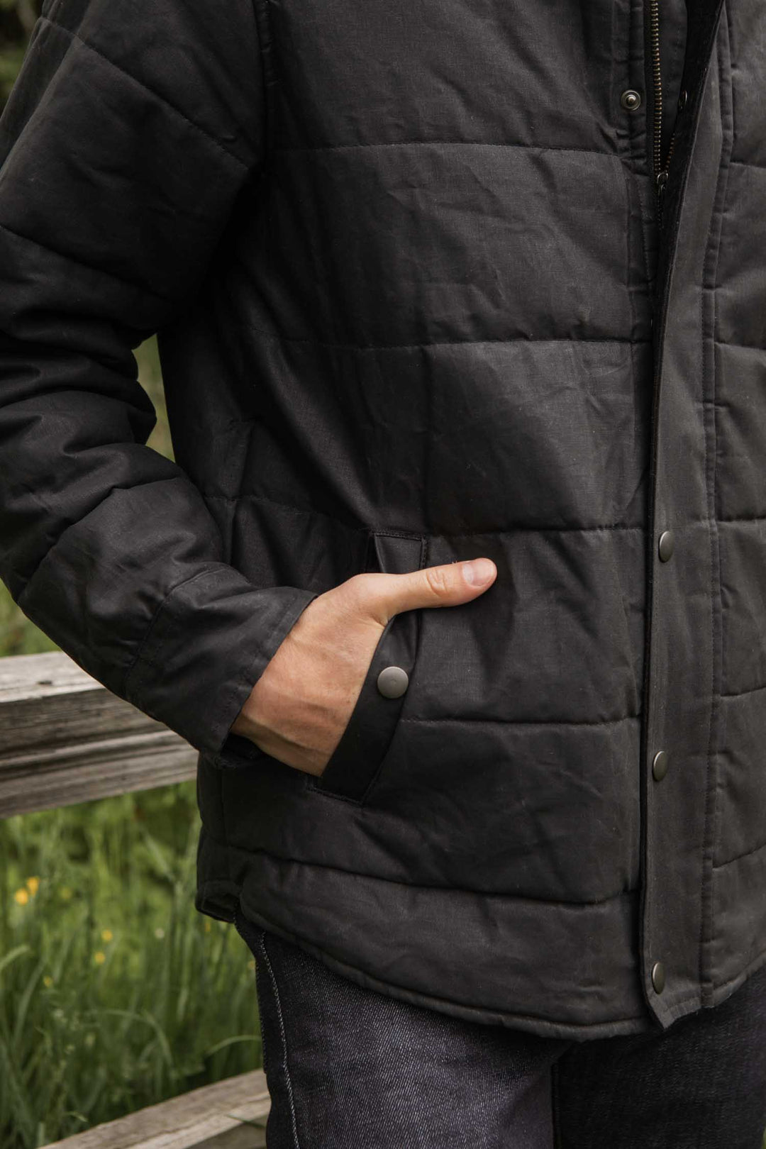 A person wearing the Bridge & Burn Holman Quilted Jacket in black waxed canvas, with their left hand in a snap-button pocket, stands outdoors by a wooden railing with greenery behind them.