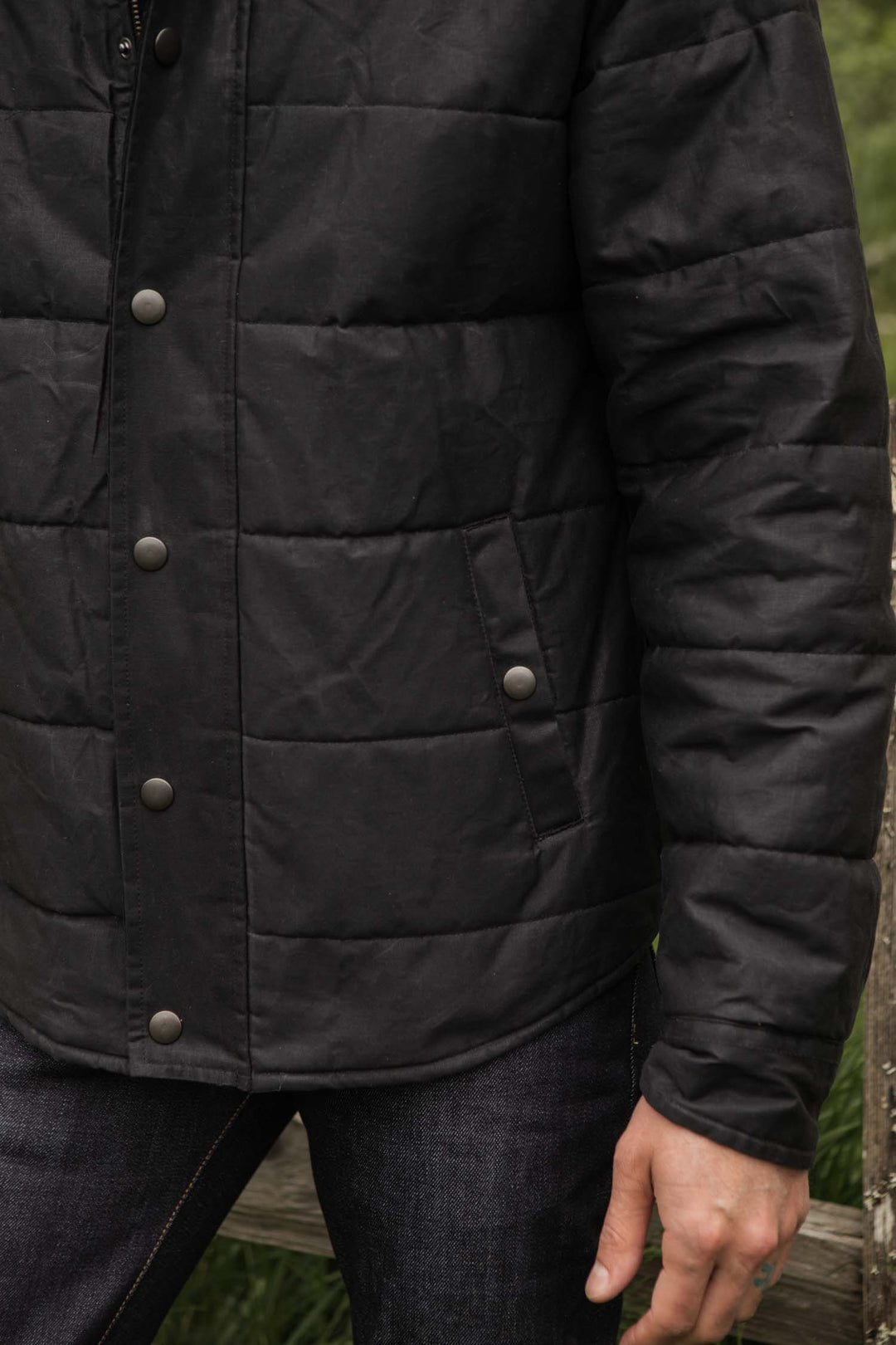 A person stands outdoors by a wooden fence, wearing the Bridge & Burn Holman Quilted Jacket in black waxed canvas. Their arm is relaxed at their side, highlighting the jacket’s texture and snap buttons.