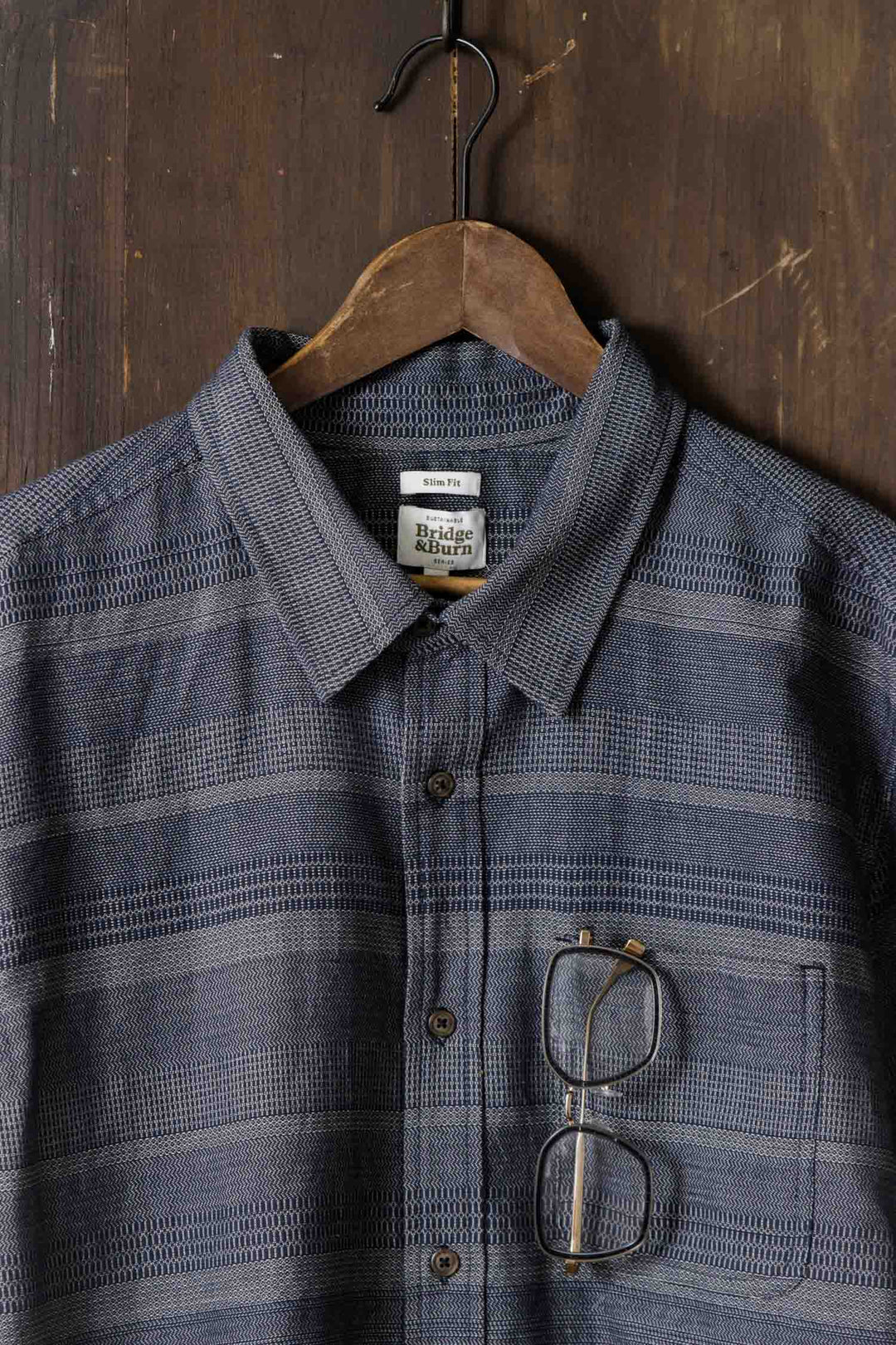 The Bridge & Burn Jude Shirt in Cascade Weave, a classic fit men's button-up with a blue pattern, hangs on a wooden hanger against a dark wood background. Eyeglasses are hooked onto the shirt’s front pocket.