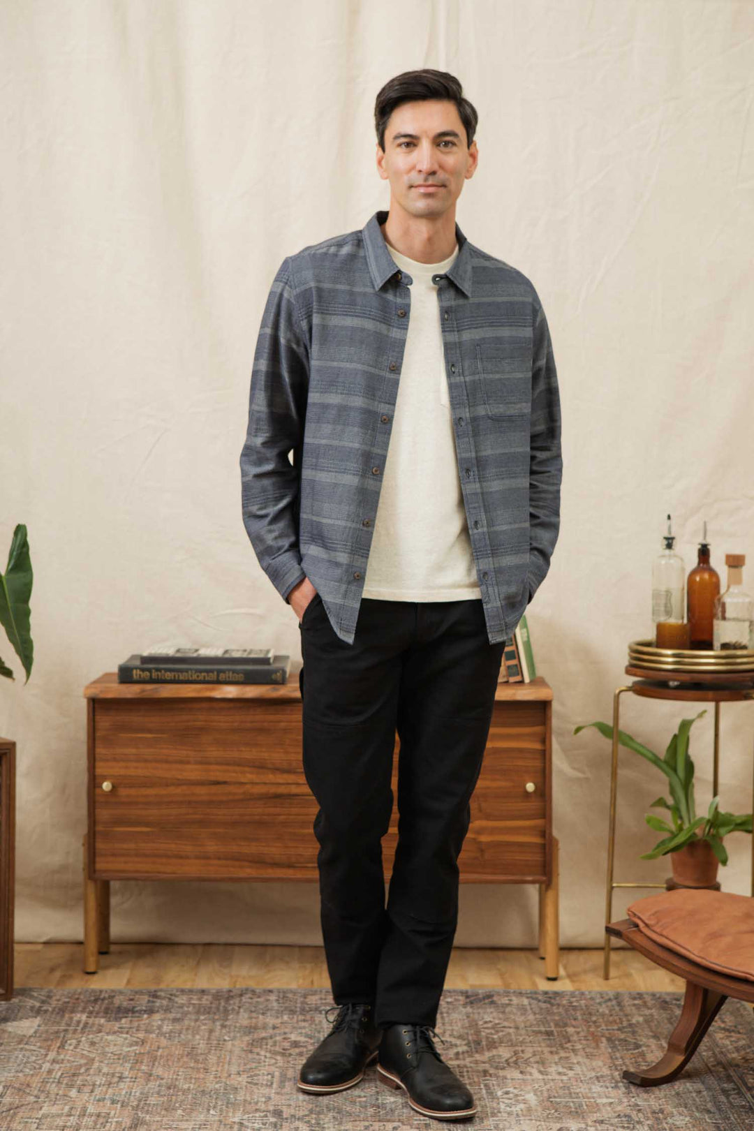A man stands indoors in the Bridge & Burn Jude Shirt / Cascade Weave—a classic fit, gray striped button-up over a white tee—paired with black pants and shoes. He’s beside a mid-century cabinet, plants, bottles, and a patterned rug.