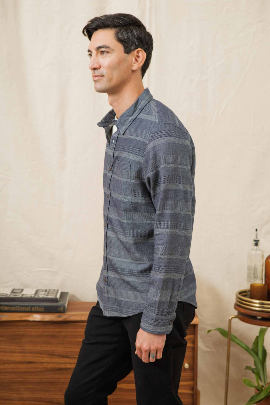 A man with short dark hair stands sideways indoors near a wooden cabinet, wearing the Bridge & Burn Jude Shirt in Cascade Weave and black pants. Decorative items and a plant are visible in the background.