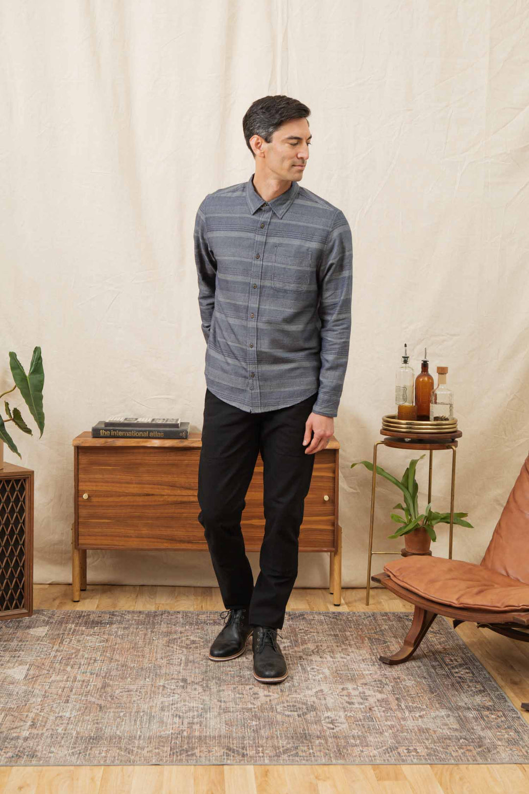 A man wearing the Bridge & Burn Jude Shirt in Cascade Weave and black pants stands indoors on a patterned rug, next to a wooden cabinet and brown leather chair, with plants decorating the background.