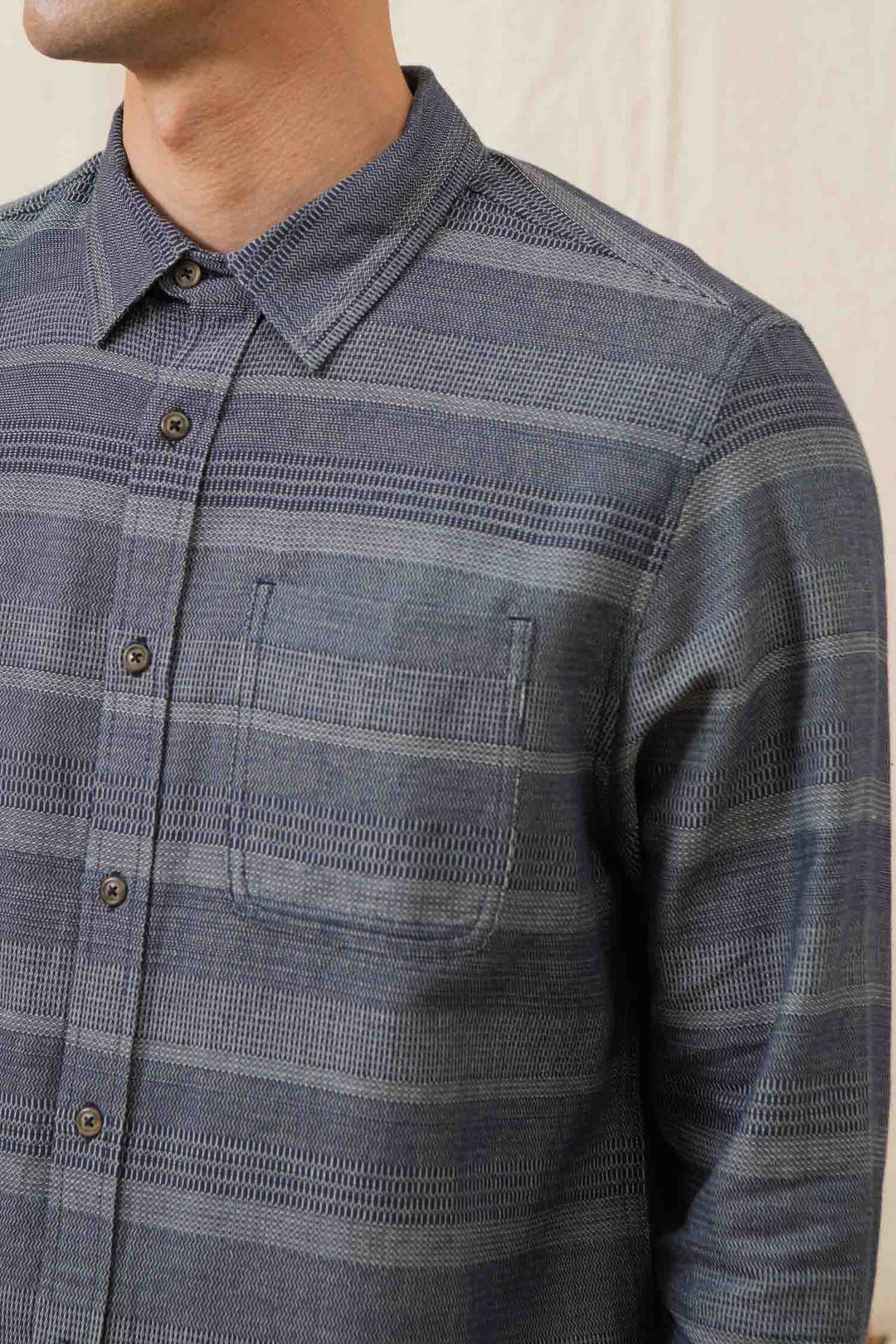 A man wearing the Bridge & Burn Jude Shirt in Cascade Weave, a blue and gray striped organic cotton button-up with a chest pocket, is shown from shoulders to mid-torso, his face partially out of frame.