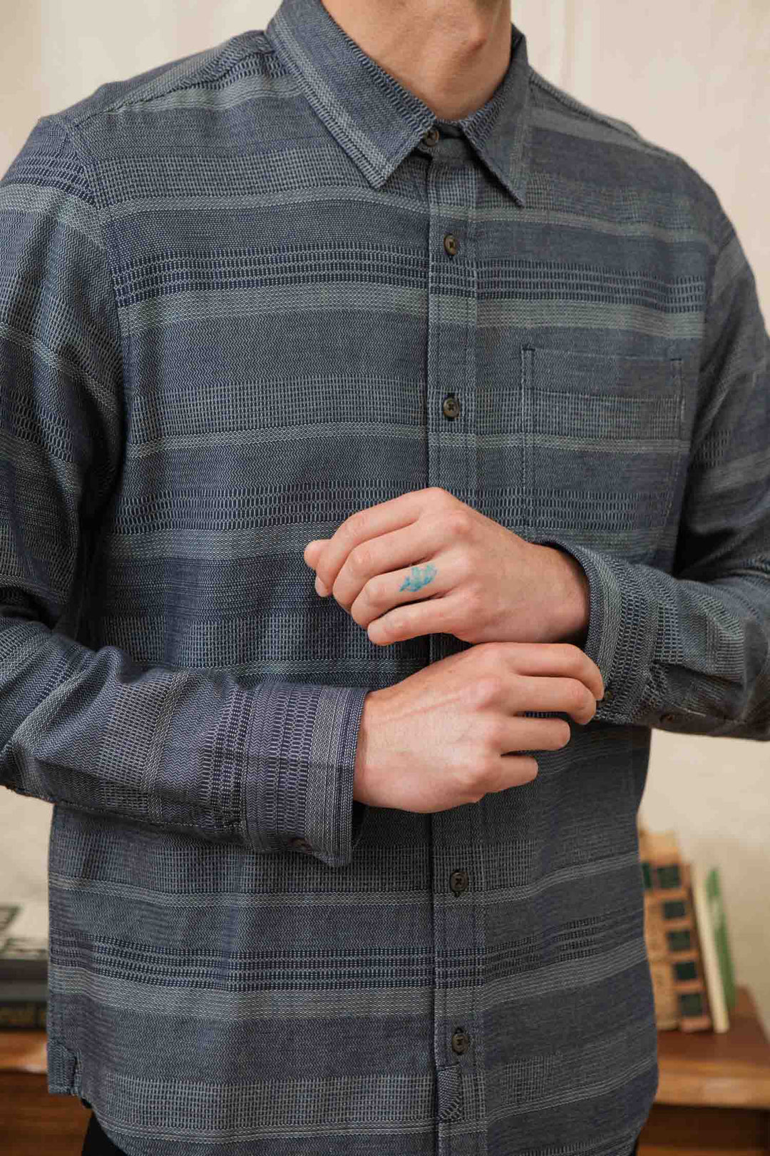 Someone wearing the Bridge & Burn Jude Shirt in Cascade Weave with a blue pattern adjusts their sleeve. A small tattoo is visible on their right ring finger, while blurred books and a beige wall fill the background. Their face is partially out of view.