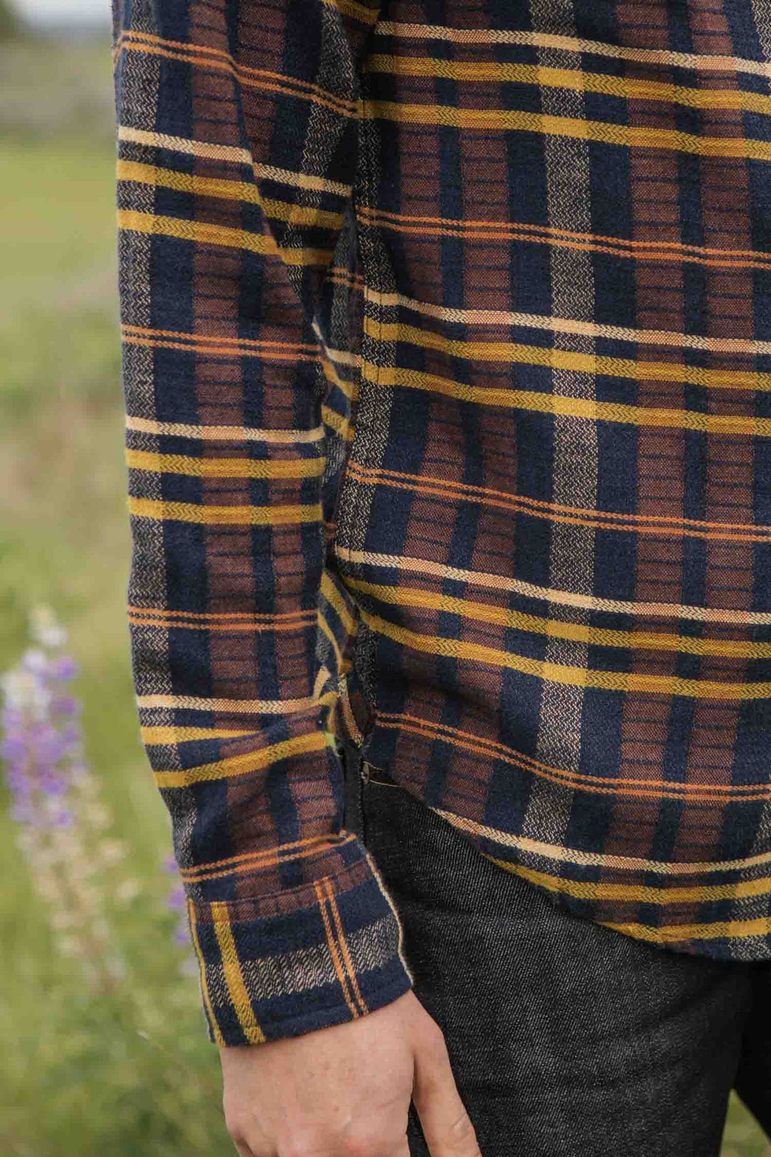 A person in the Bridge & Burn Larson Shirt / Desert Dawn Plaid, a classic fit long-sleeve flannel in brown, yellow, and navy, stands outdoors on grass with their arm relaxed by their side. Dark pants and blurred flowers are partially visible.