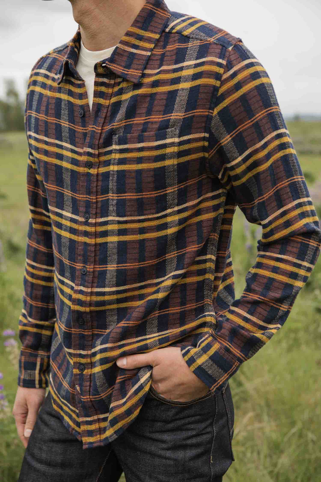 A person stands outdoors in a grassy area, wearing the Bridge & Burn Larson Shirt in Desert Dawn Plaid over a white undershirt, with one hand in their jeans pocket. The person's face is not fully visible.