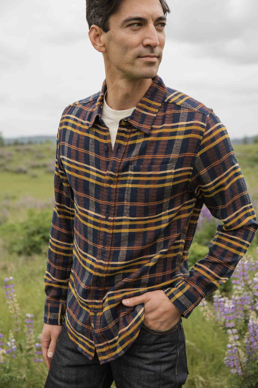 A man wearing the Bridge & Burn Larson Shirt in Desert Dawn Plaid and jeans stands in a grassy field with purple wildflowers, looking to the side. The sky is cloudy and the background is softly blurred.