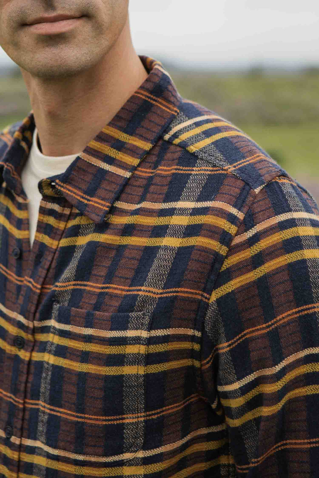 A close-up of a man wearing the Bridge & Burn Larson Shirt in Desert Dawn Plaid, a classic fit organic cotton flannel with navy, mustard, and brown stripes. Only his lower face and upper body are visible against a blurred green background.