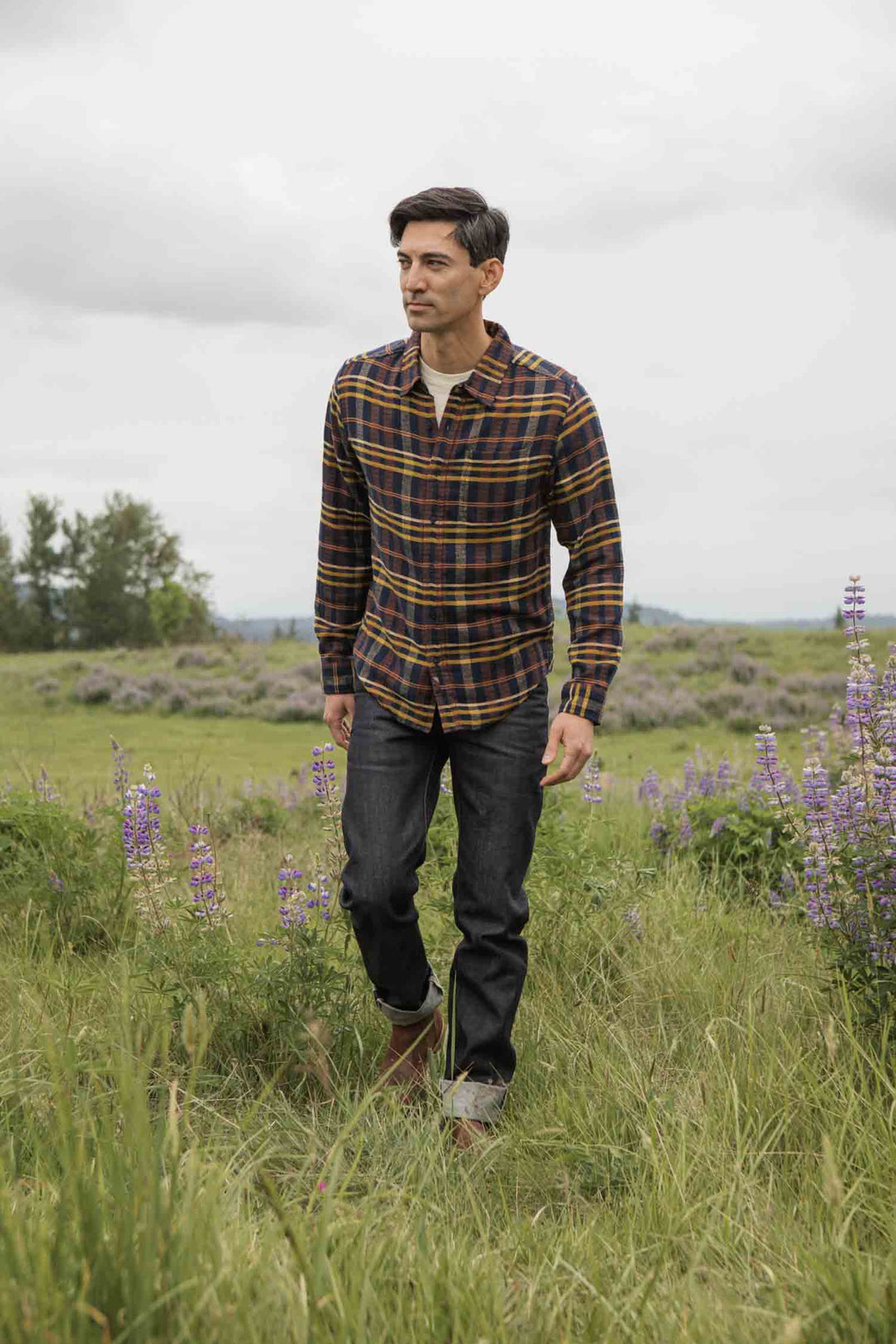 A man wearing the Bridge & Burn Larson Shirt in Desert Dawn Plaid and jeans walks outdoors through a grassy field with purple wildflowers beneath a cloudy sky.