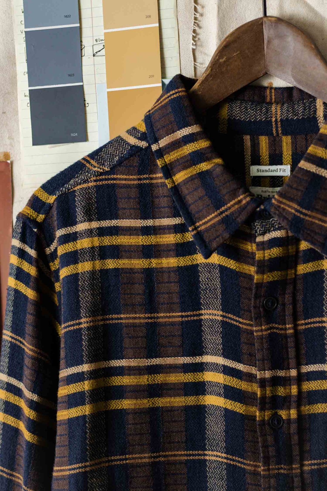 A close-up of the Bridge & Burn Larson Shirt in Desert Dawn Plaid, a classic fit button-up flannel with navy, brown, and yellow stripes on a wooden hanger; blue, tan, and brown paint swatches are visible in the background.