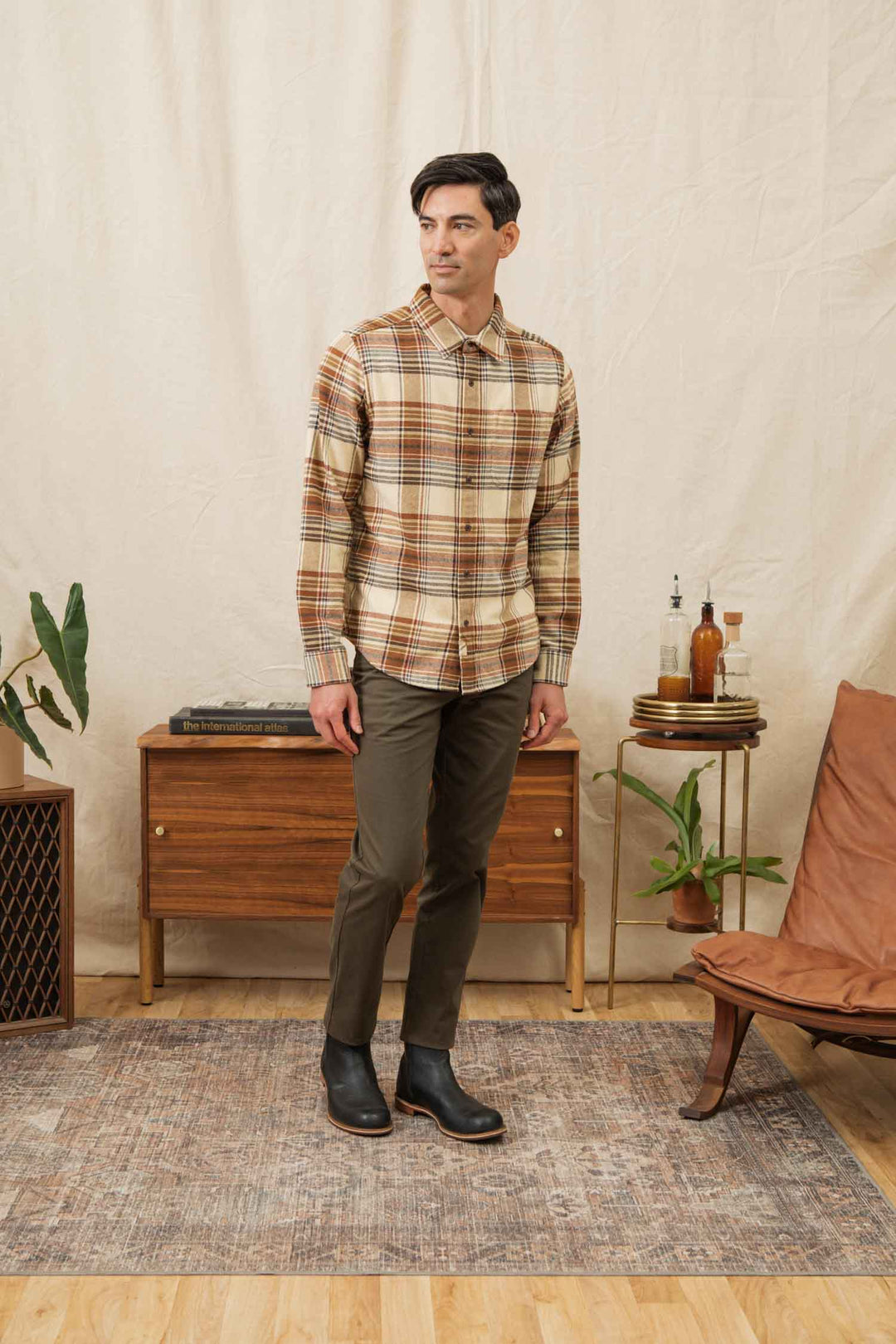 A man stands on a rug indoors, wearing the Bridge & Burn Larson Shirt in Latte Plaid with olive pants and black boots. Behind him are a wooden cabinet with books and bottles, two potted plants, and a brown leather chair.