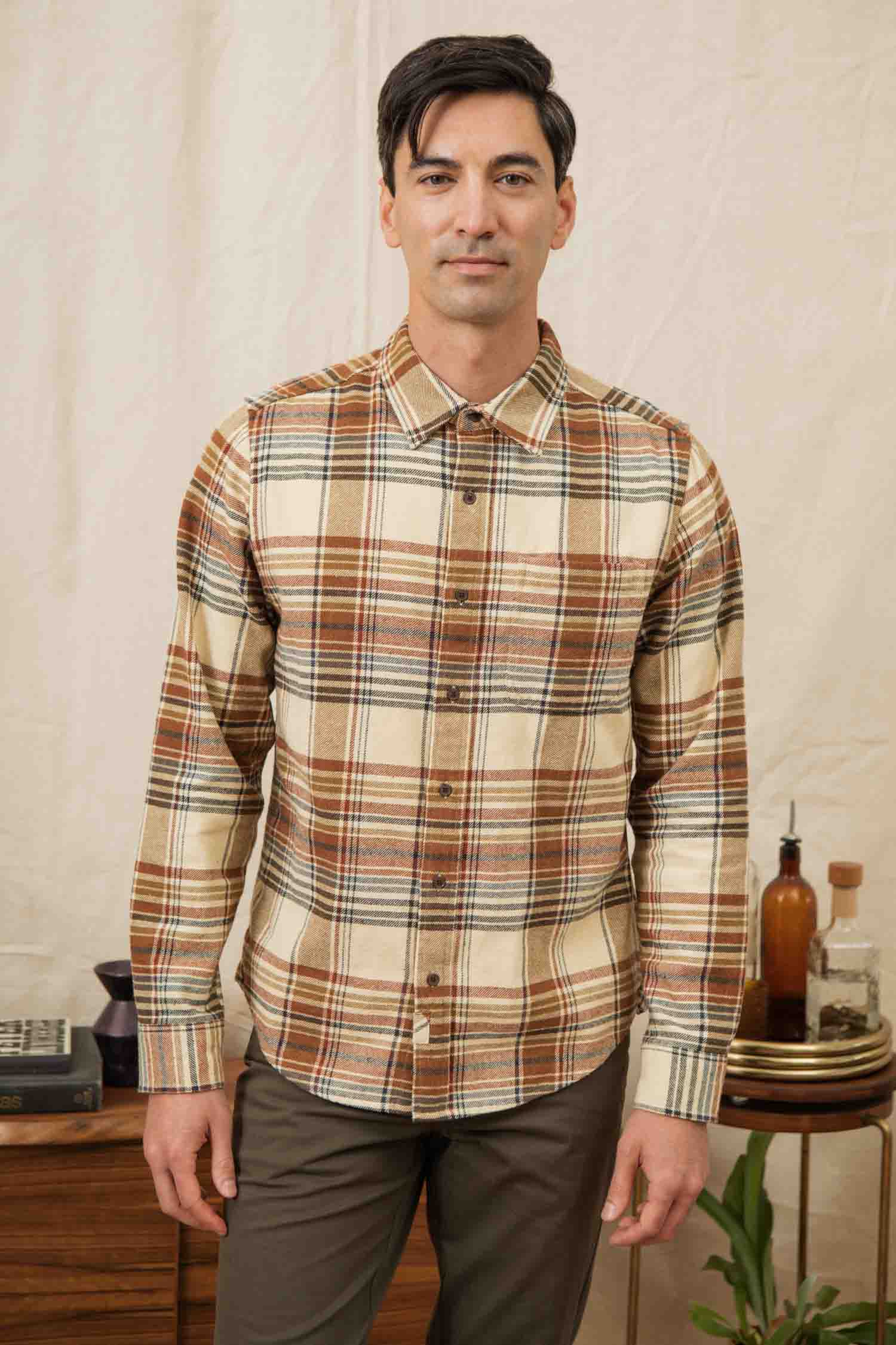 A man with dark hair wears the Bridge & Burn Larson Shirt in Latte Plaid and brown pants, standing indoors in front of a light curtain with wooden furniture and decor behind him.