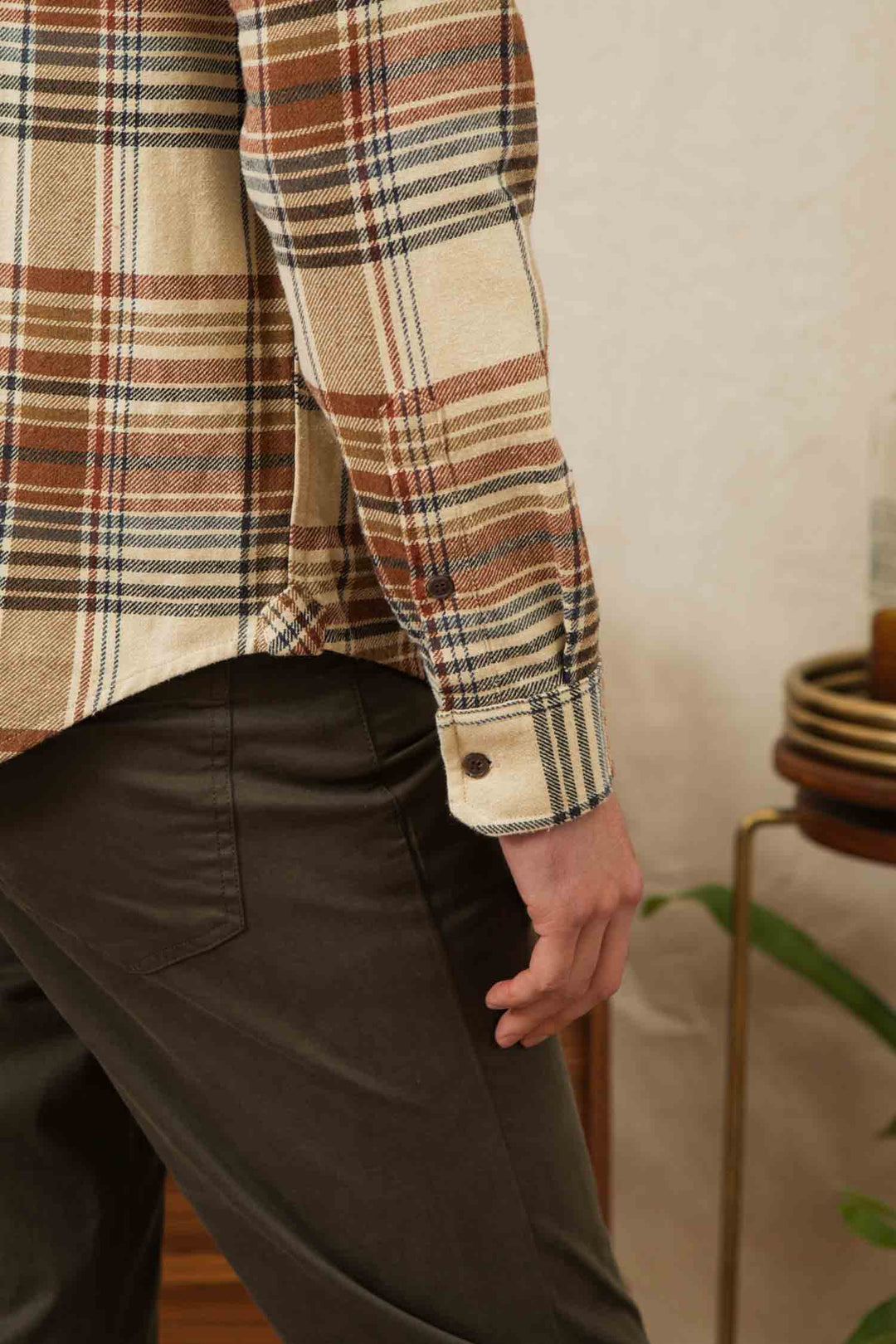 A person wears the Bridge & Burn Larson Shirt in Latte Plaid with dark pants, standing indoors with their arm relaxed. A small table holding stacked plates and a bottle is visible in the background.