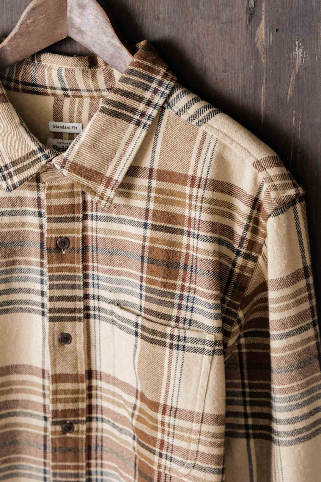 The Bridge & Burn Larson Shirt in Latte Plaid—a beige, brown, and cream brushed flannel classic-fit button-up with collar and chest pocket—is crafted from recycled cotton for sustainability. Shown hanging on a wooden hanger against rustic wood.
