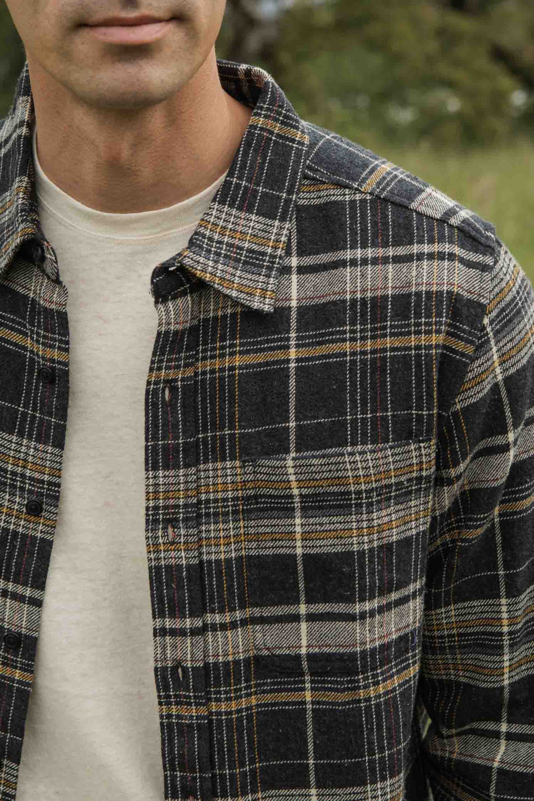 A person stands outdoors, wearing the Bridge & Burn Larson Shirt in Volcanic Plaid over a cream T-shirt; only the lower face and upper torso are visible, with blurred greenery in the background.