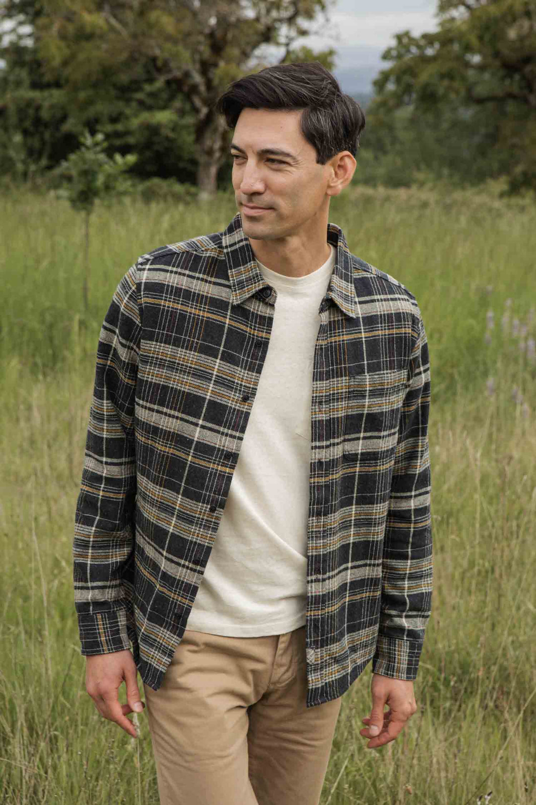 A man stands in a grassy field, smiling slightly as he looks left. He wears the Bridge & Burn Larson Shirt in Volcanic Plaid over a cream t-shirt and beige pants. Trees and greenery are visible in the background.