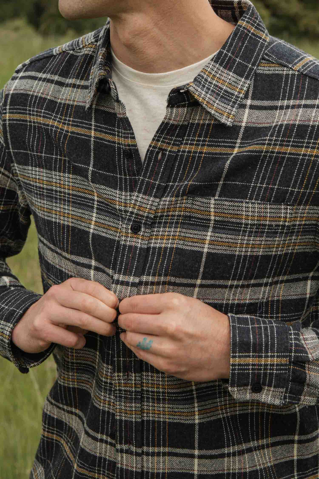 A person outdoors is buttoning up the Bridge & Burn Larson Shirt in Volcanic Plaid, a classic fit brushed flannel, worn over a white T-shirt. Only their torso and lower face are visible, with grass in the background.