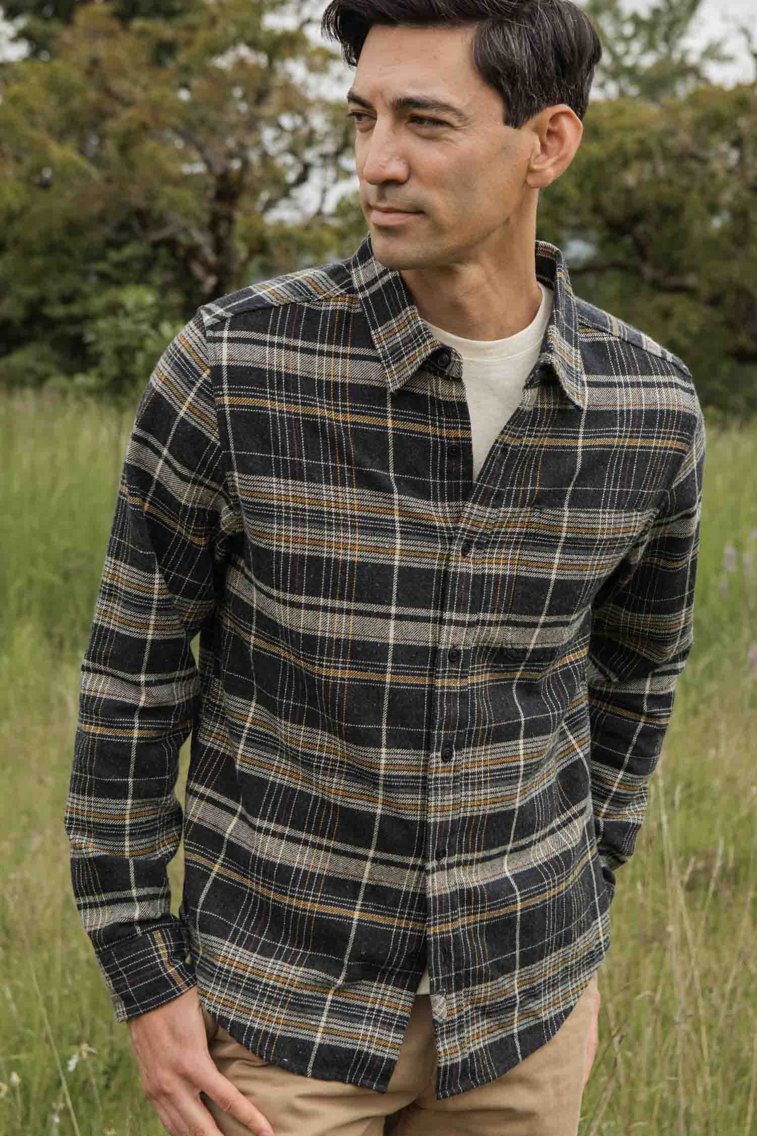 A man stands in a grassy field wearing the Bridge & Burn Larson Shirt in Volcanic Plaid over a light T-shirt and tan pants, looking to the side with trees behind him.