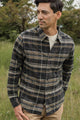 A man stands in a grassy field wearing the Bridge & Burn Larson Shirt in Volcanic Plaid over a light T-shirt and tan pants, looking to the side with trees behind him.