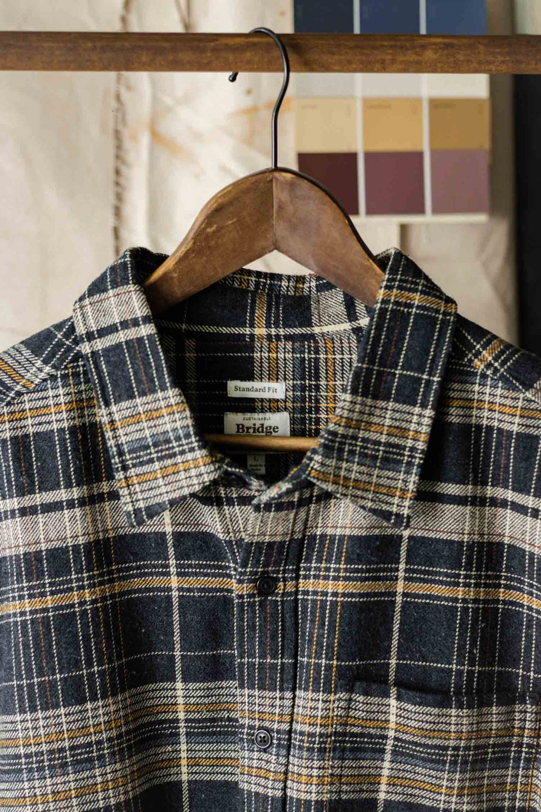 The Bridge & Burn Larson Shirt in Volcanic Plaid—a brushed flannel button-up in black, white, and yellow—hangs on a wooden hanger with a blurred paint swatch chart and off-white fabric in the background.