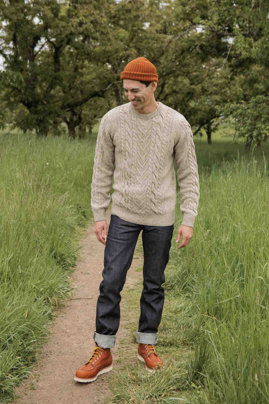 A man in the Bridge & Burn Ludlow Sweater in Flecked Oatmeal, dark jeans, brown boots, and an orange beanie stands on a grassy path smiling, with trees behind him.