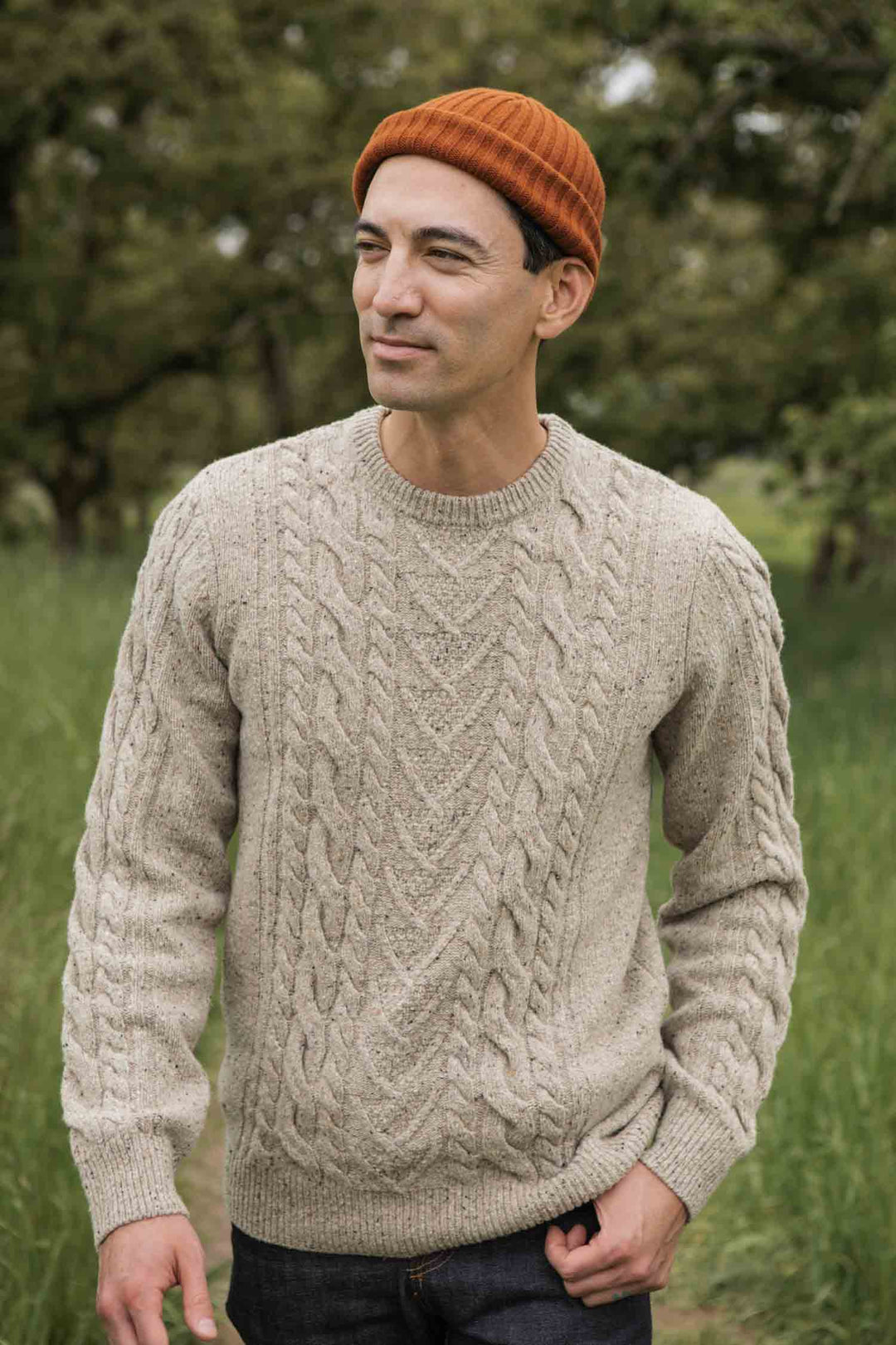 A man stands outdoors in a grassy area with trees, smiling gently and looking to the side, wearing the Bridge & Burn Ludlow Sweater in Flecked Oatmeal and an orange beanie.