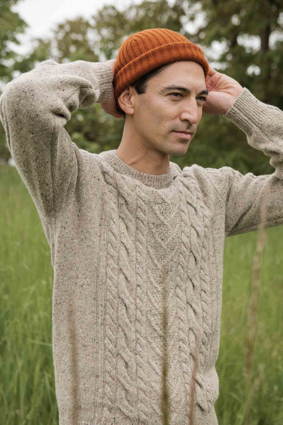 A person wears the Bridge & Burn Ludlow Sweater in Flecked Oatmeal (beige, cable-knit, RWS-certified wool) and an orange knit beanie, standing in a grassy field with trees, hands behind their head.