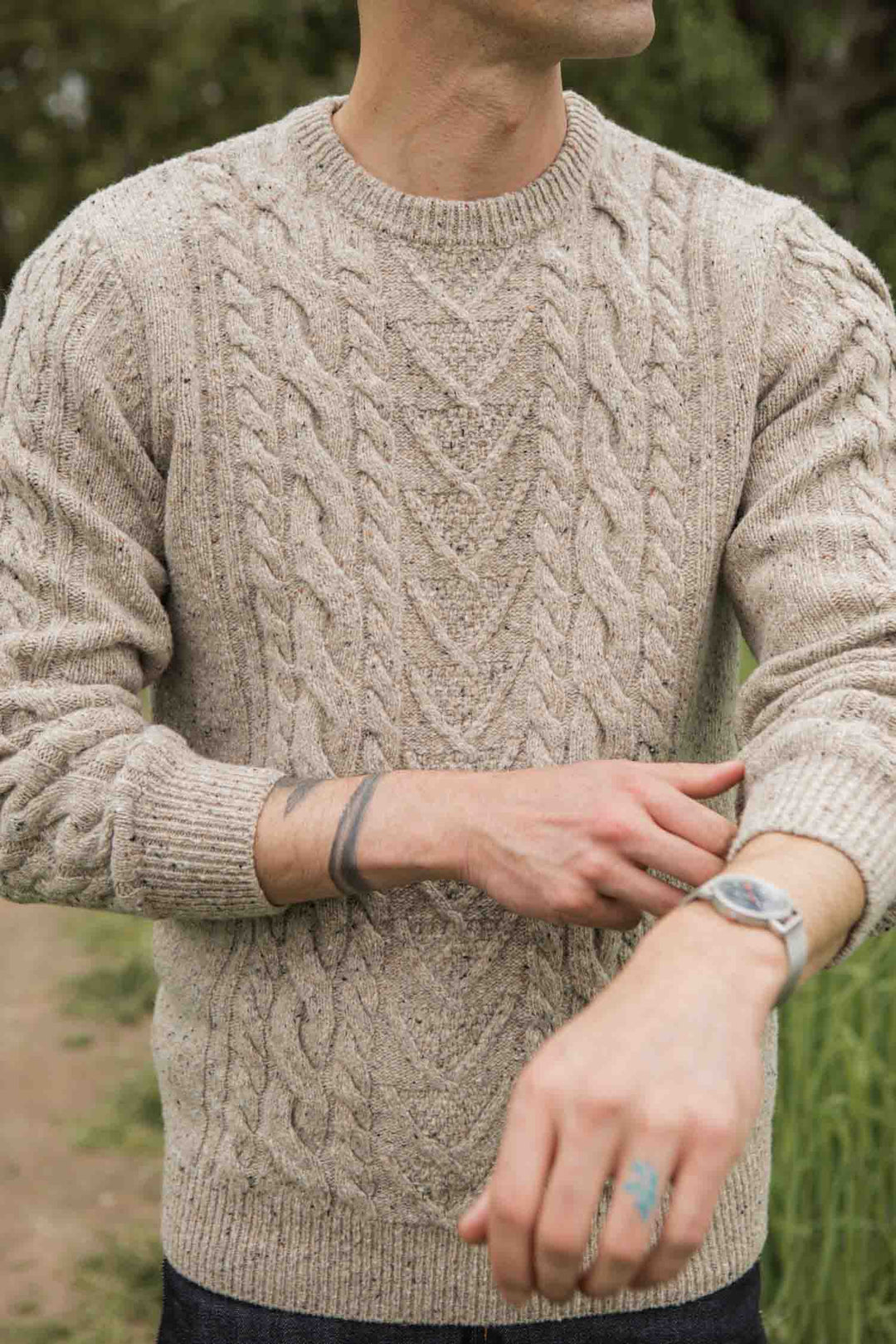 Wearing a Bridge & Burn Ludlow Sweater in Flecked Oatmeal, dark jeans, bracelet, and wristwatch, a person stands outdoors amid greenery. Their face is partially out of frame.