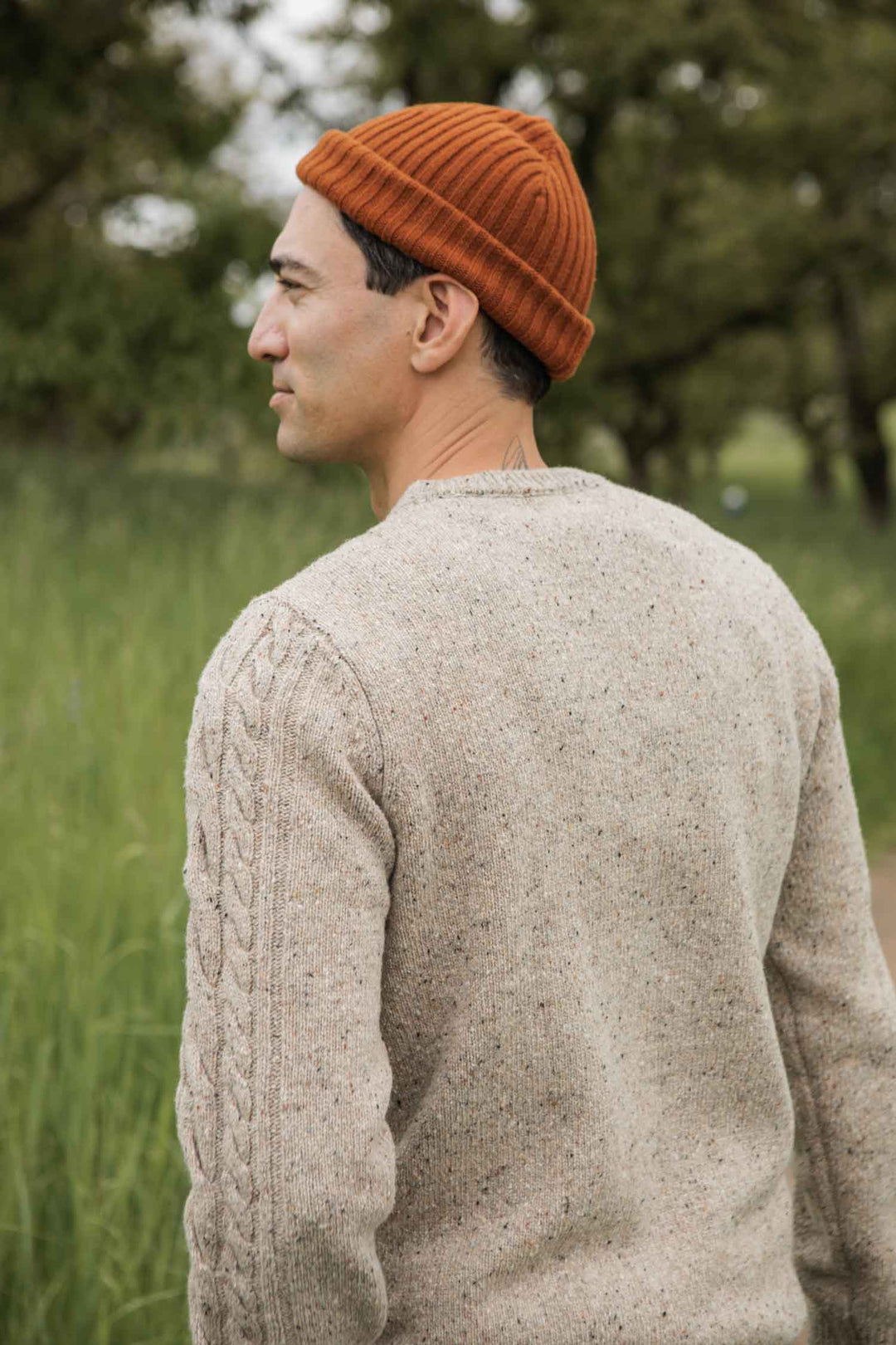 A man in the Bridge & Burn Ludlow Sweater in Flecked Oatmeal and an orange knit beanie stands outdoors with tall green grass and blurred trees behind him, facing away to the left.