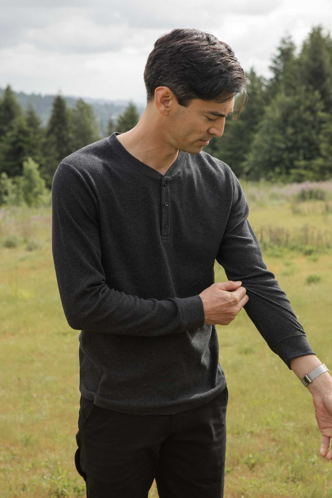 A man stands in a grassy field with trees behind him, looking down as he adjusts the sleeve of his Bridge & Burn Men's Recycled Waffle Henley in Charcoal Melange.