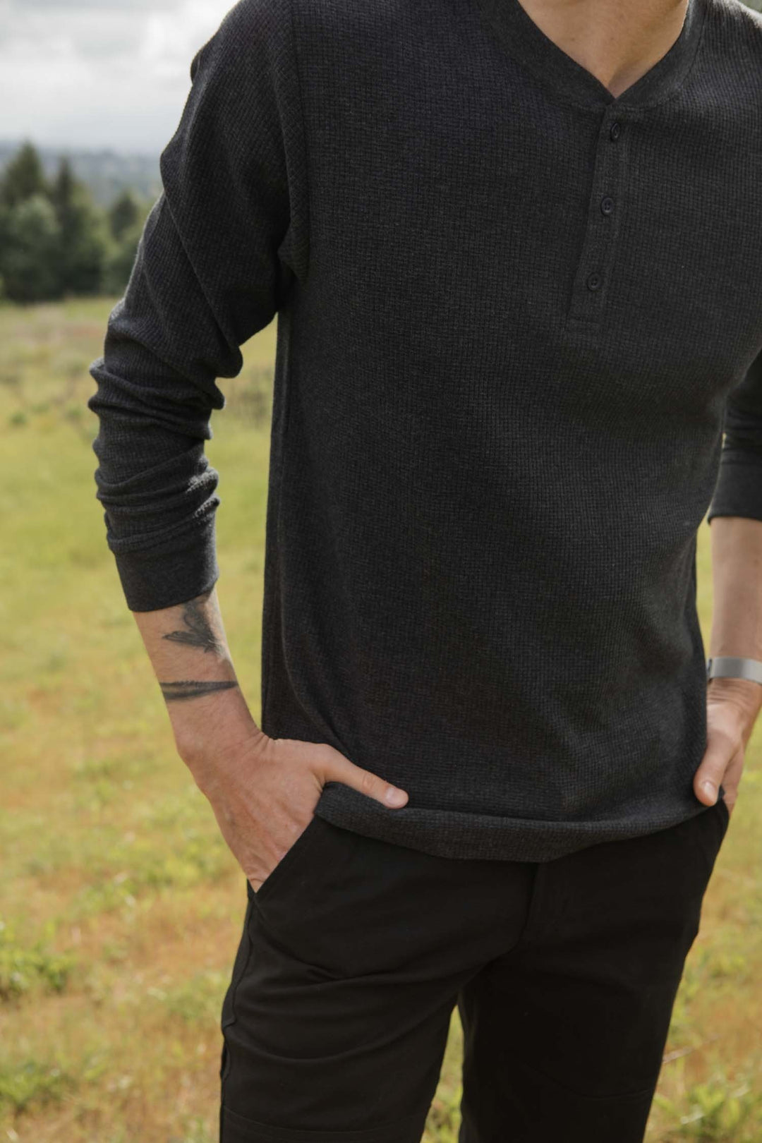 A man stands outdoors with hands in pockets, wearing the Bridge & Burn Men's Recycled Waffle Henley in Charcoal Melange and black pants. His tattooed forearm is visible. The casual, sustainable look is set against greenery.