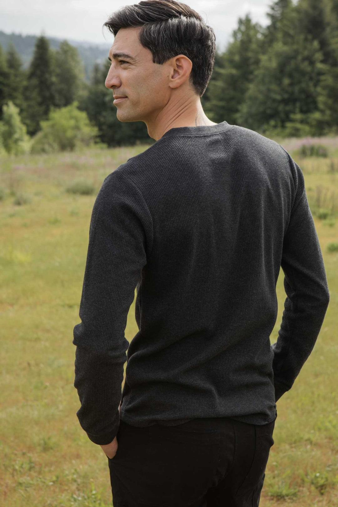 A man with short dark hair stands in a grassy field, looking over his shoulder. He wears the Bridge & Burn Men's Recycled Waffle Henley in Charcoal Melange. Trees fill the background and his hands rest in his pockets.