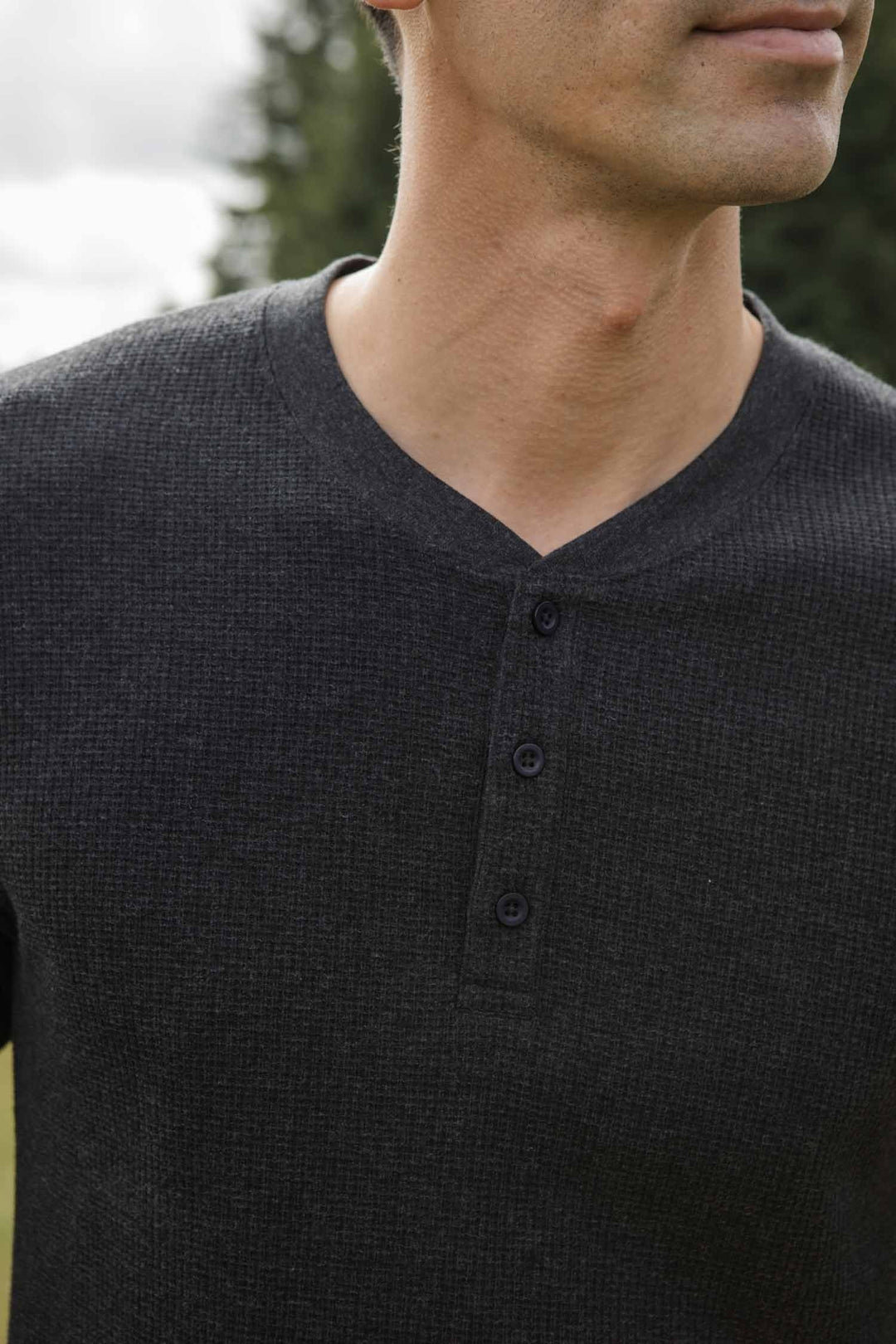 A man stands outdoors among blurred trees, wearing Bridge & Burn's Men's Recycled Waffle Henley in Charcoal Melange—a dark gray, three-button shirt crafted from recycled fibers. His face is partially out of frame.