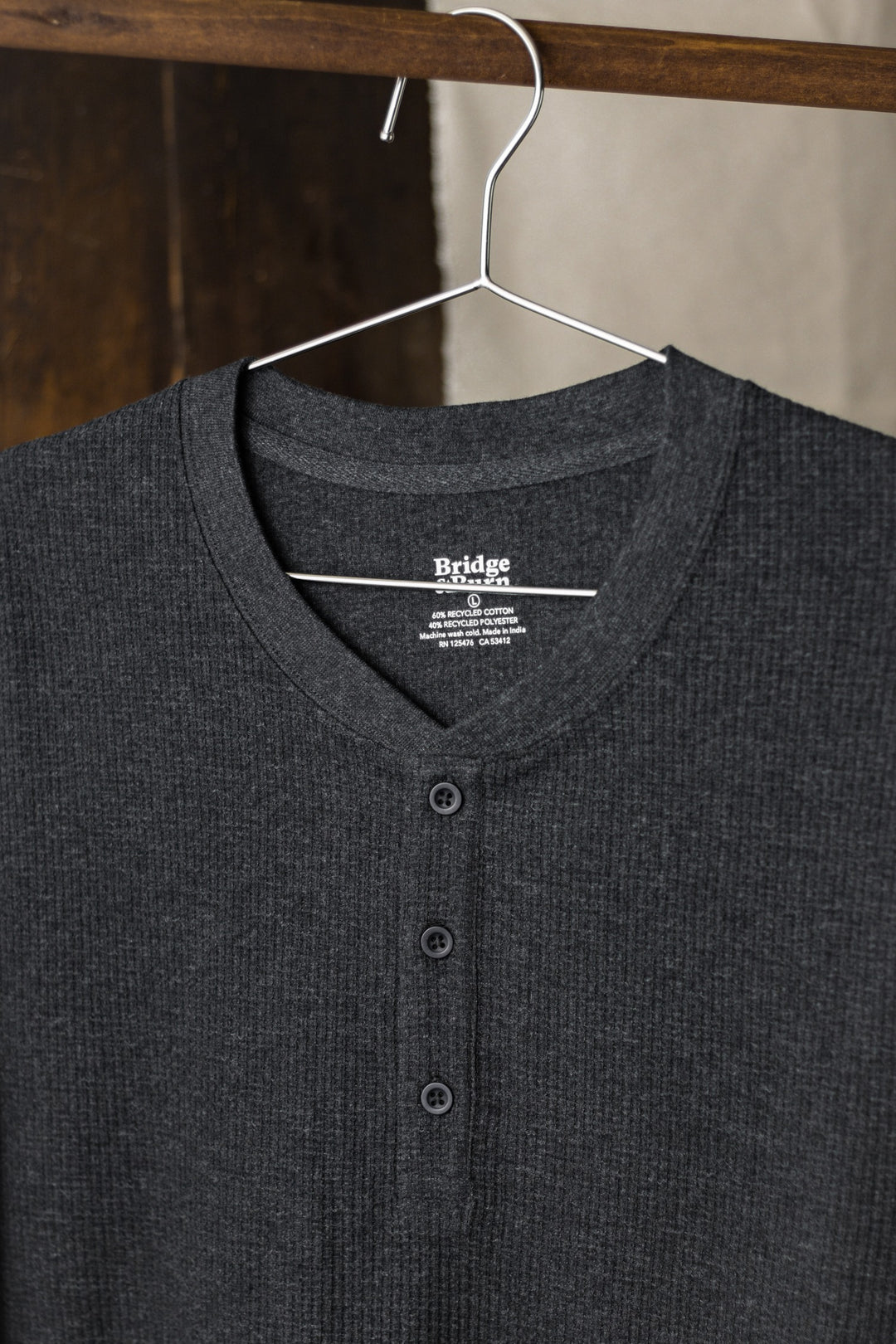 A charcoal melange men's recycled waffle henley with long sleeves and three buttons by Bridge & Burn hangs on a thin metal hanger against a wooden background.