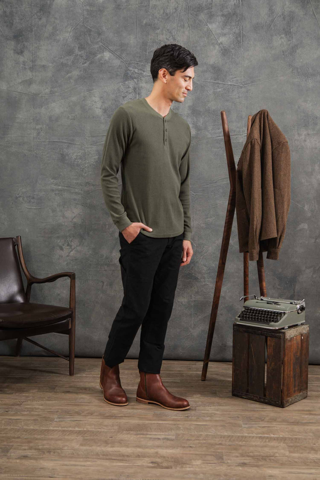A man stands indoors on a wooden floor, wearing Bridge & Burn's Men's Recycled Waffle Henley in Forest Melange, black pants, and brown boots. Beside him are a chair, coat rack with a brown jacket, and a vintage typewriter on a crate.