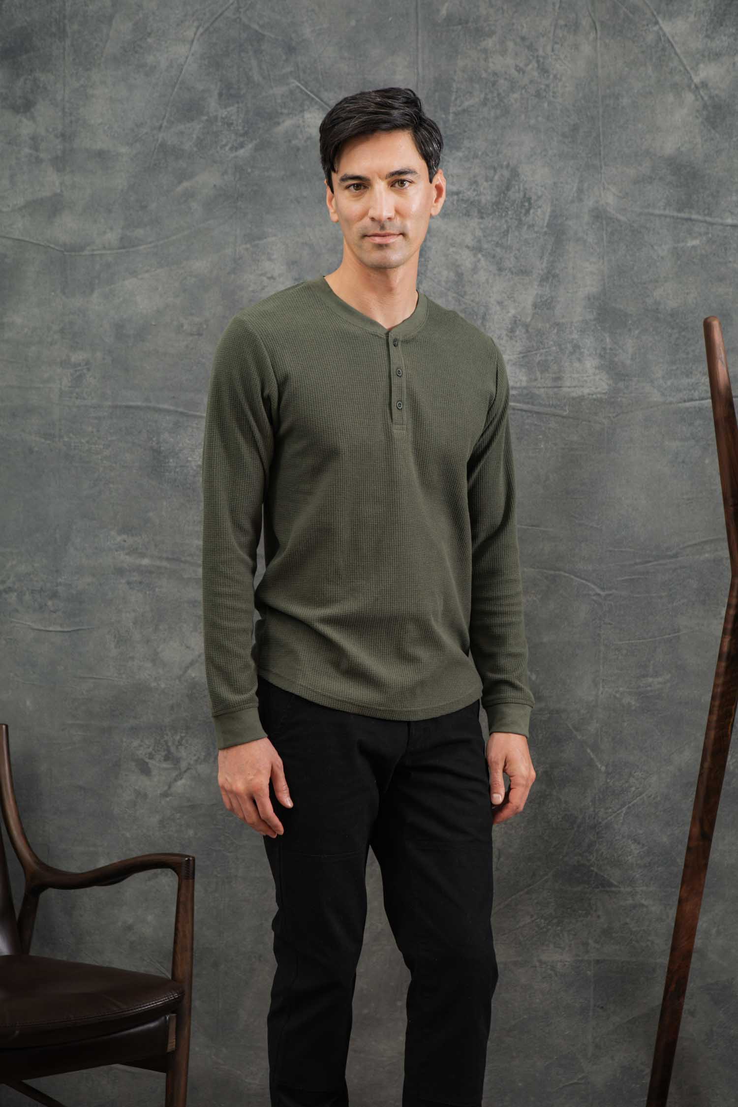 A man with short dark hair stands indoors against a gray textured backdrop, wearing Bridge & Burn’s Men's Recycled Waffle Henley in Forest Melange and black pants, with wooden chairs partially visible on either side.