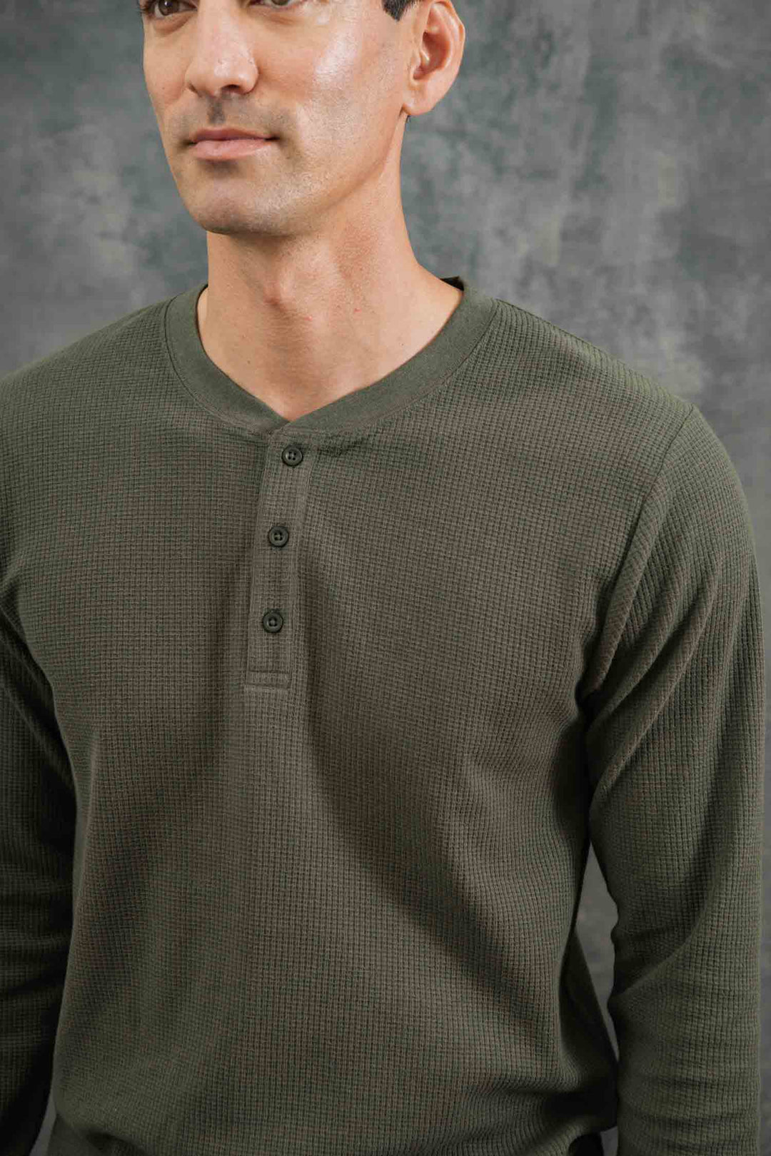 A man stands before a textured gray background, partially visible, wearing the Bridge & Burn Men's Recycled Waffle Henley in Forest Melange.