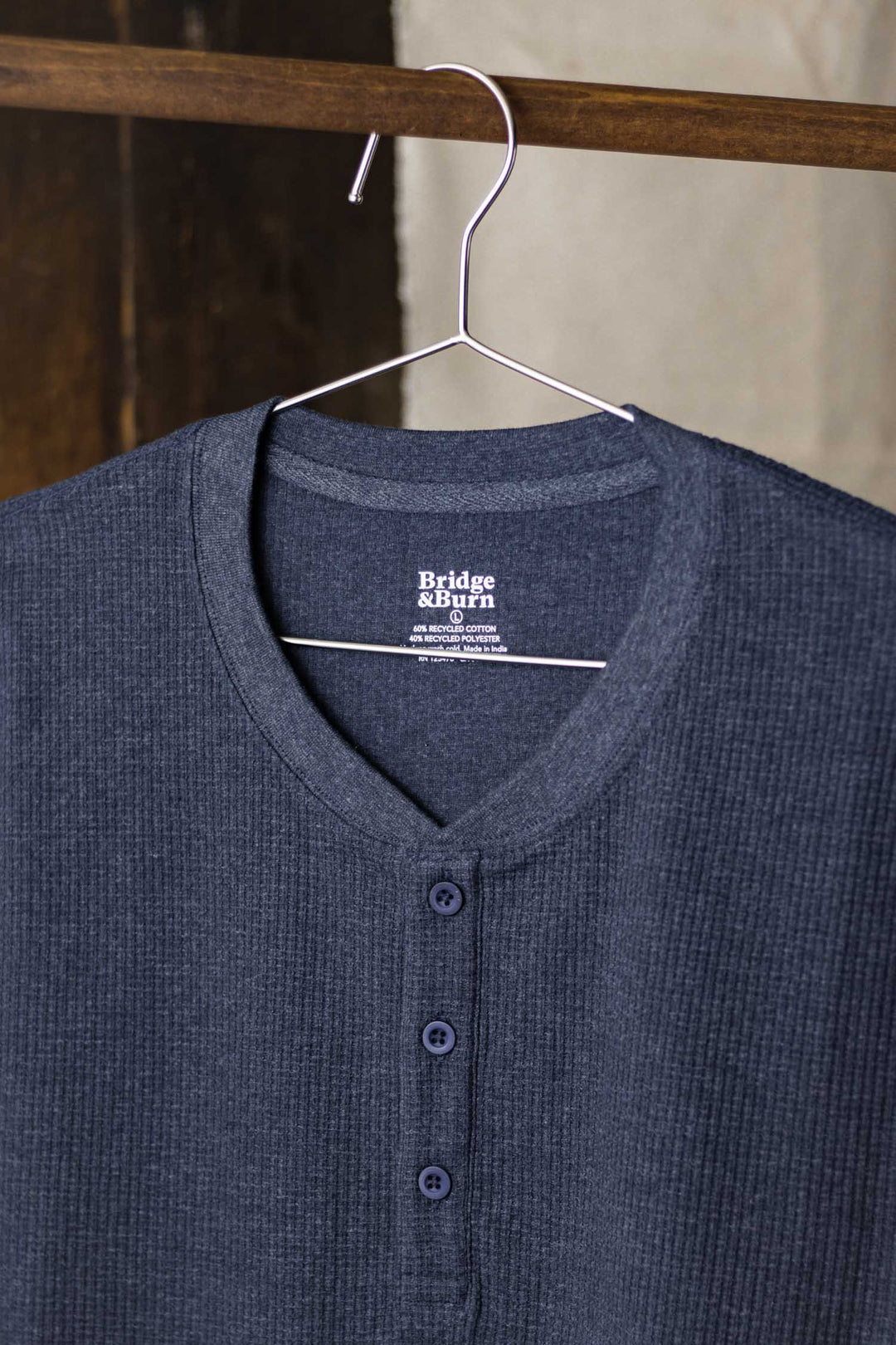 A Men's Recycled Waffle Henley in Sapphire Melange by Bridge & Burn hangs on a wire hanger against a wooden beam and light wall. Three buttons detail the front, showcasing its stylish and sustainable waffle-knit design.