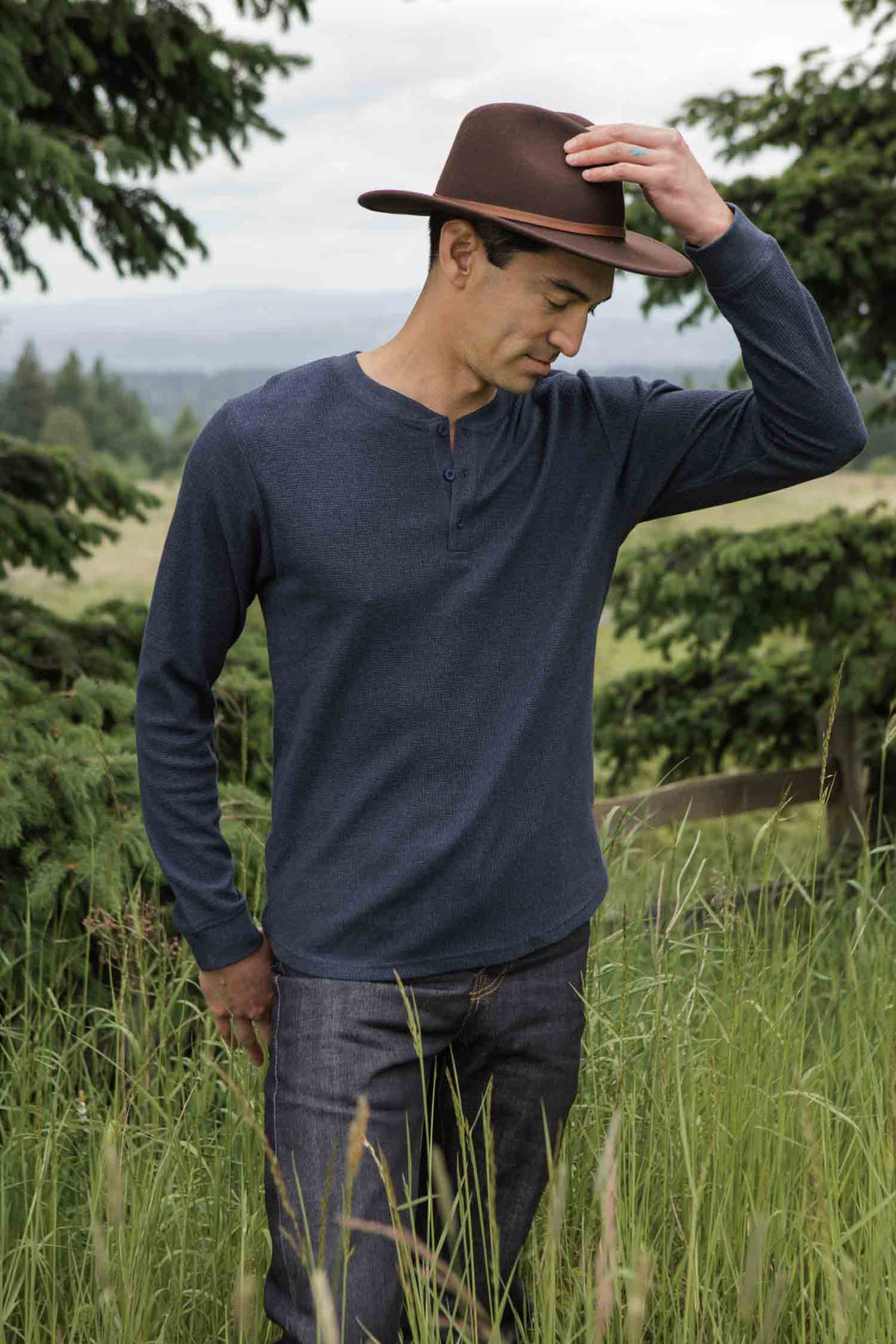 A man in tall grass adjusts his brown hat, wearing Bridge & Burn’s Men's Recycled Waffle Henley in Sapphire Melange and jeans, with pine trees and a cloudy sky behind him.