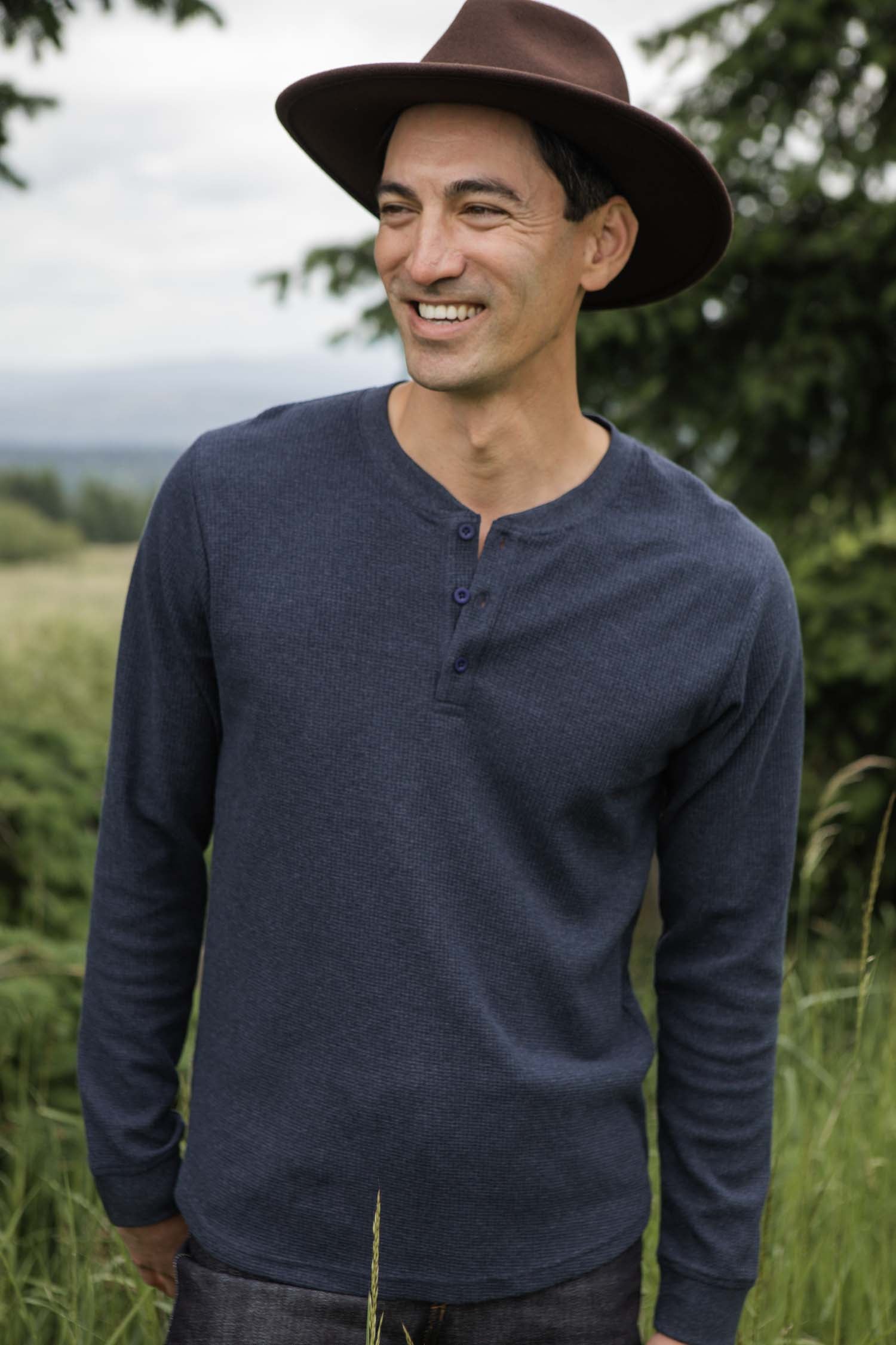 A smiling man wears the Bridge & Burn Men's Recycled Waffle Henley in Sapphire Melange and a brown wide-brimmed hat, standing outdoors amid grass, trees, and distant hills.