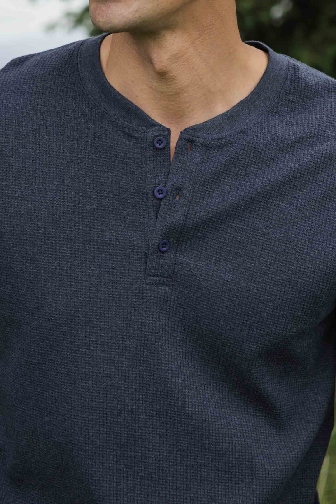 A person wearing the Bridge & Burn Men's Recycled Waffle Henley in Sapphire Melange, featuring long sleeves and a three-button collar. Only their chest, neck, and part of the face are shown against a blurred outdoor background.