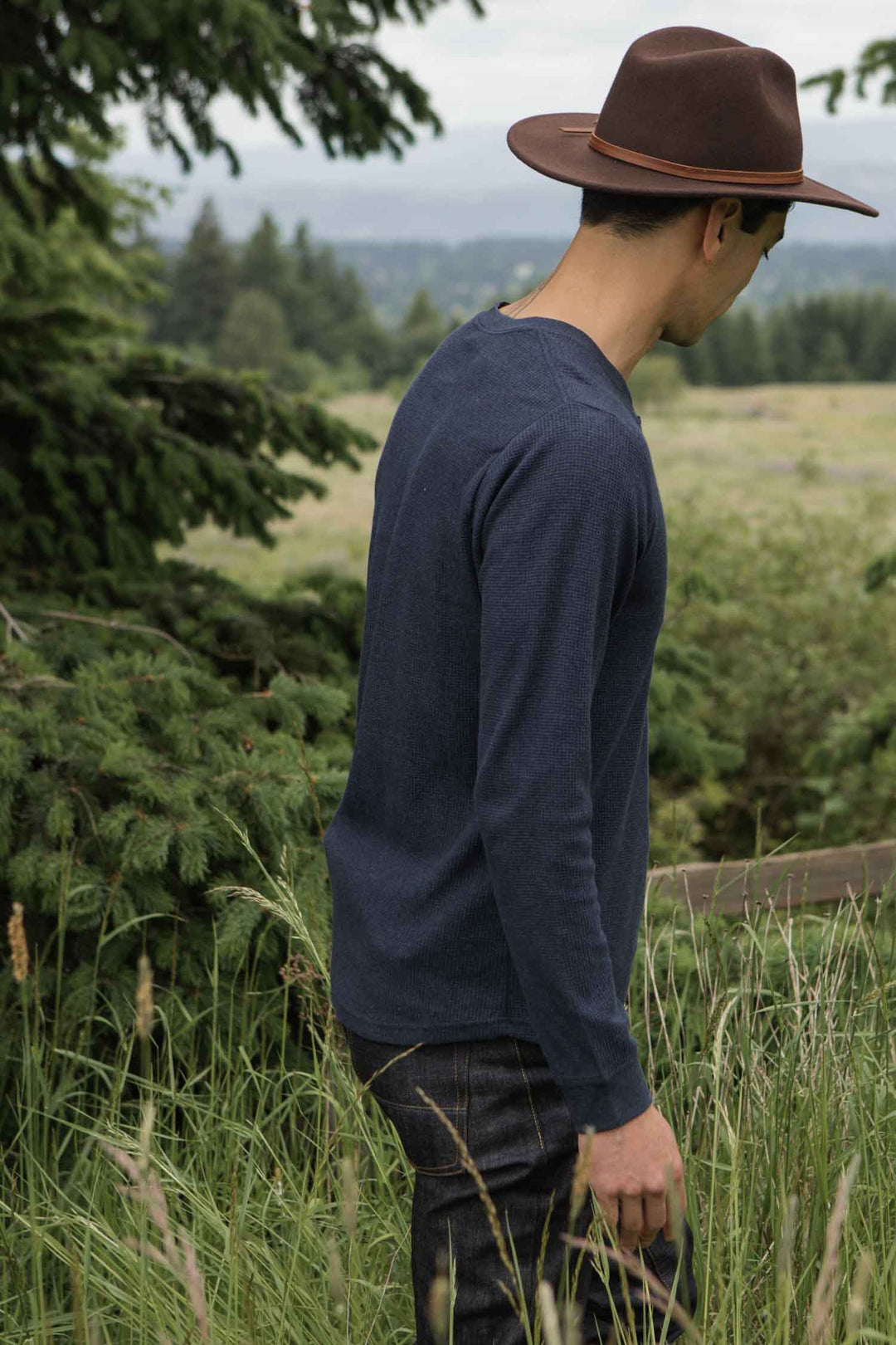 A person in a Bridge & Burn Men's Recycled Waffle Henley in Sapphire Melange and a brown hat stands in tall grass, facing away, with trees and an open field behind them.