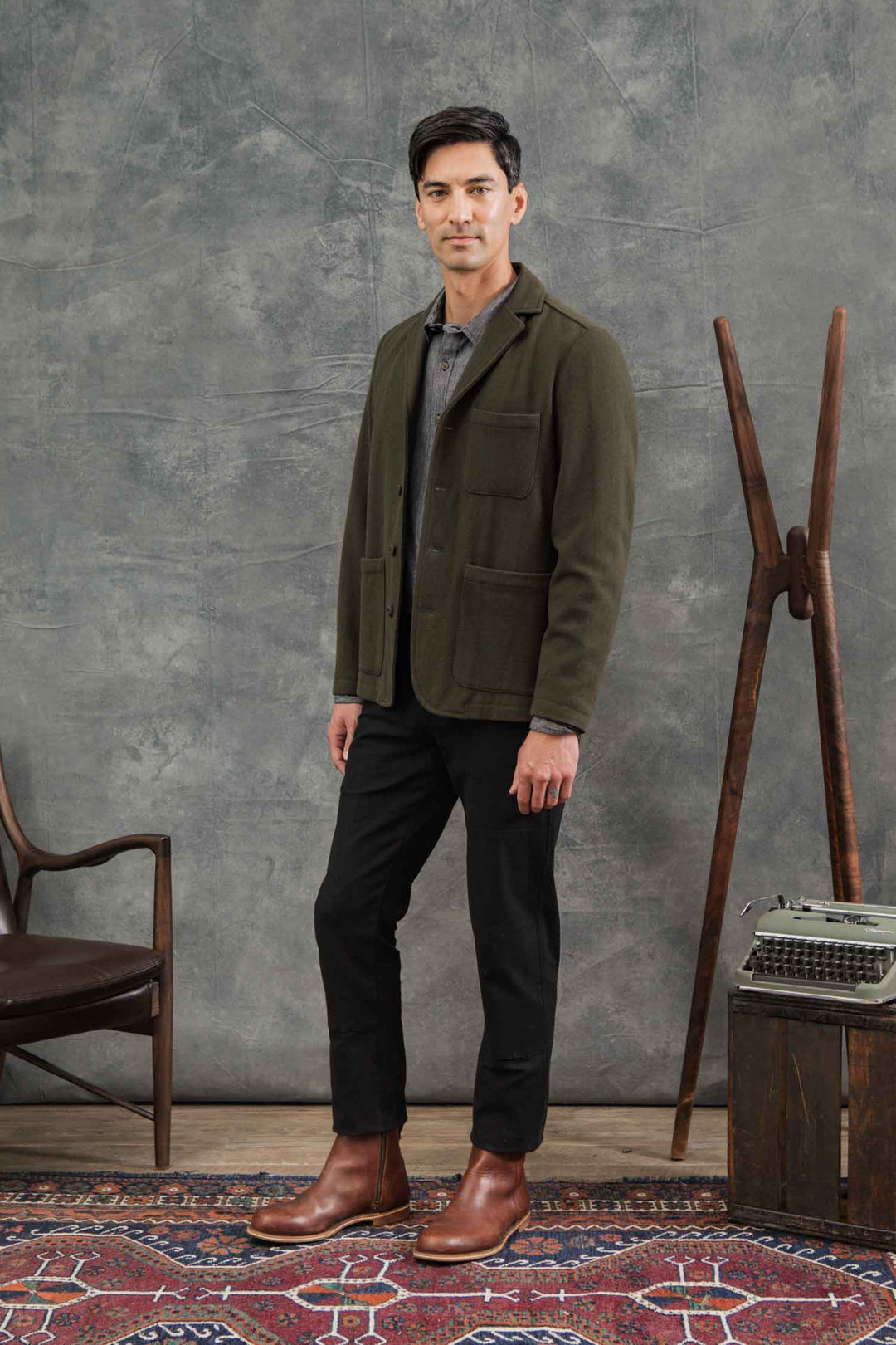 A man stands indoors on a patterned rug, wearing the Bridge & Burn Nate Chore Blazer in Basque Green Wool over a gray shirt, black pants, and brown boots. Behind him are a chair, mid-century coat rack, and typewriter atop a wooden box against a gray backdrop.