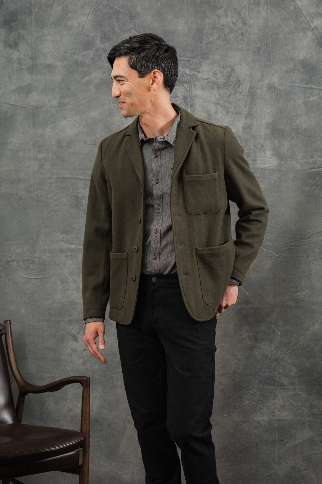 A man stands beside a wooden chair, wearing the Bridge & Burn Nate Chore Blazer in Basque Green Wool over a gray shirt and black pants. He faces left, smiling against a gray textured background.
