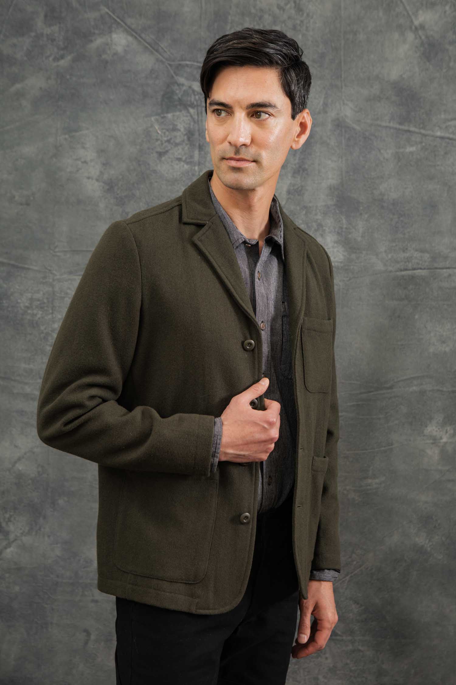 A man with short dark hair models the Bridge & Burn Nate Chore Blazer in Basque Green Wool over a gray shirt, posing against a textured gray background and looking to the side while adjusting his jacket.