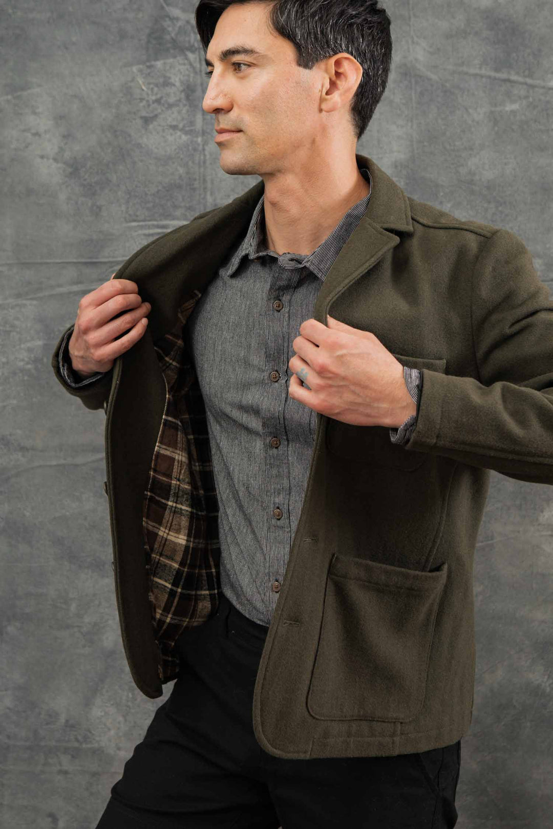 A man in a gray button-up and Basque Green Wool Nate Chore Blazer by Bridge & Burn holds one side open to show its brown plaid lining, standing against a textured gray background and looking to his left.