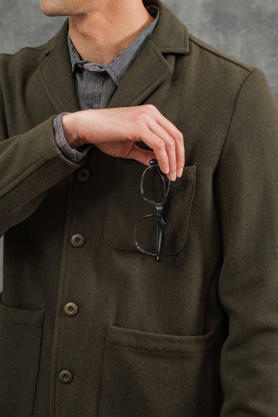 A man in a Bridge & Burn Nate Chore Blazer in Basque Green Wool wears a striped shirt and holds black eyeglasses by his chest pocket, his face partially out of frame against a gray background.