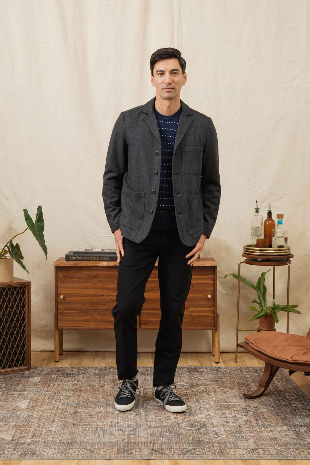 A man stands indoors on a patterned rug, wearing the Bridge & Burn Nate Chore Blazer in Onyx Wool, paired with dark pants and black sneakers. He is in front of a wooden console table with plants and bottles, set against a beige backdrop.