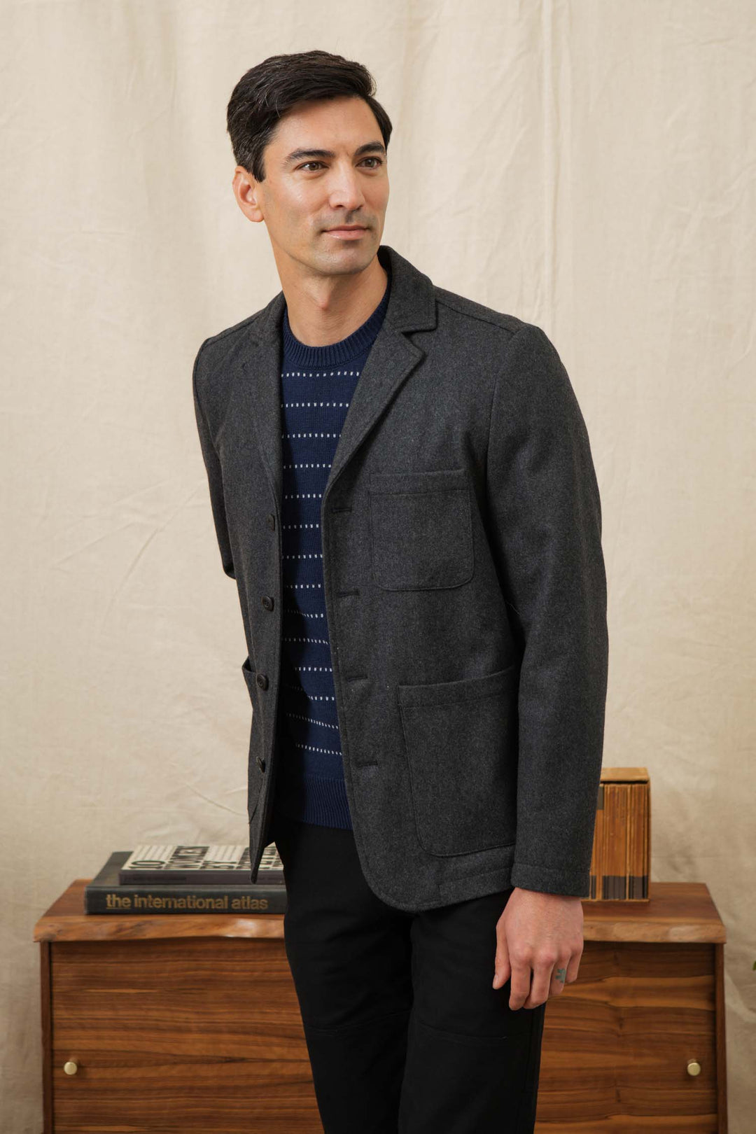 A man with dark hair wears the Bridge & Burn Nate Chore Blazer in Onyx Wool over a navy patterned sweater and black pants, standing indoors by a beige curtain next to a wooden dresser with books and a decorative box.